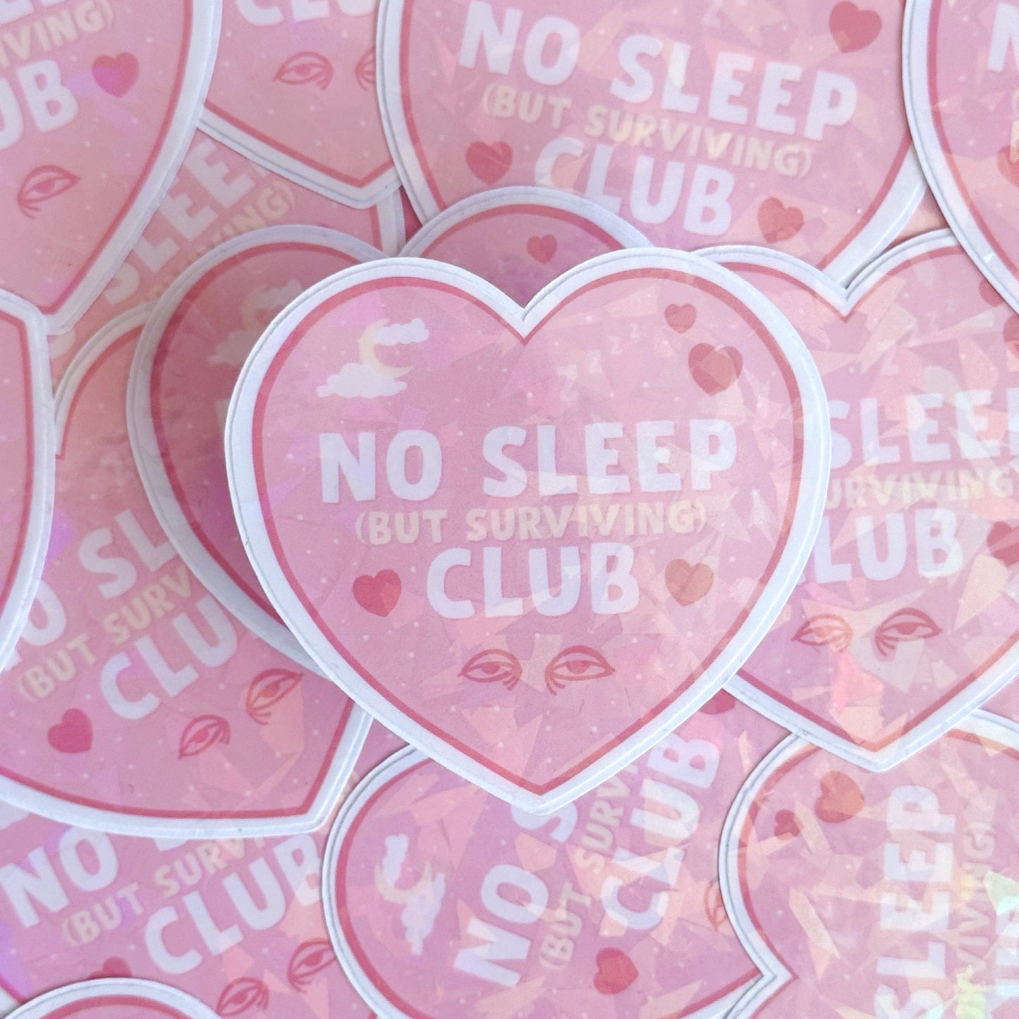 A pink holographic sticker, in the shape of a heart, with the words 'No sleep (but surviving) club' in the centre. Around the text are little doodles, like tired eyes, hearts and a moon behind clouds.