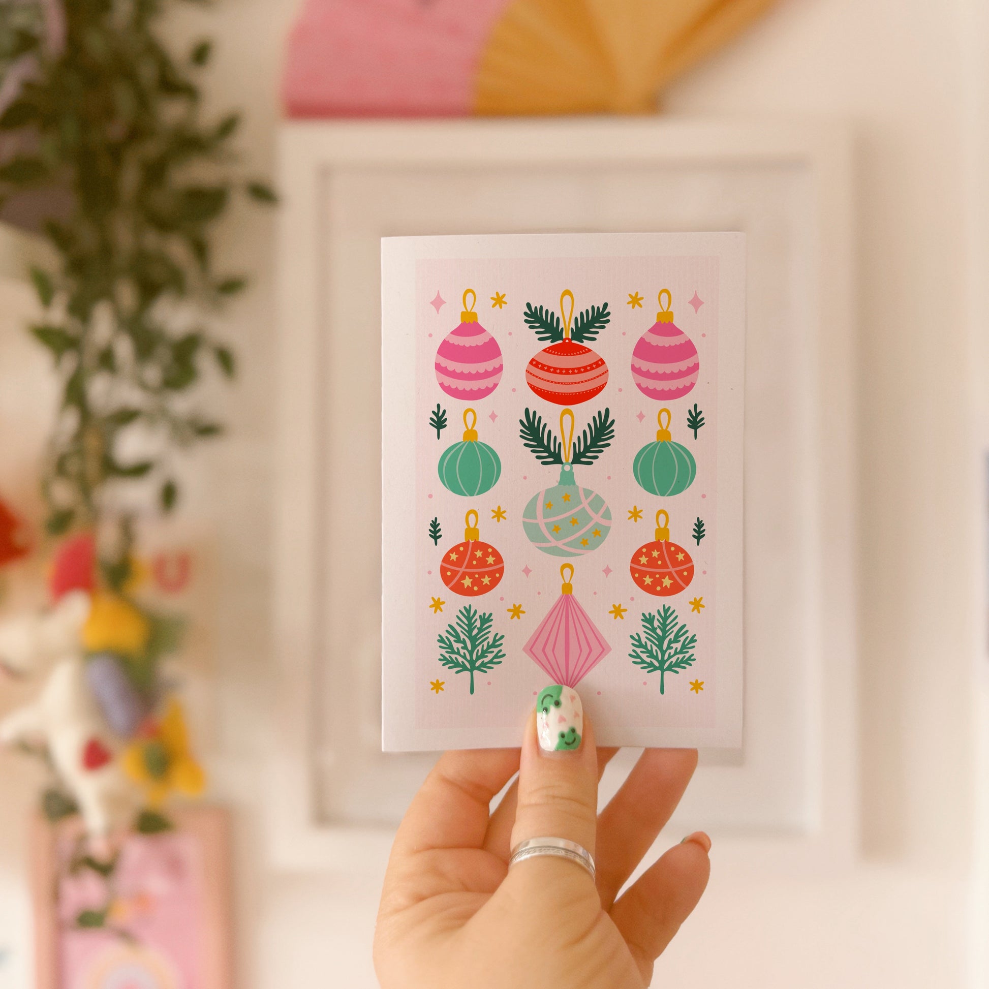 Hand holding a card with colorful Christmas ornament designs against a blurred indoor background.