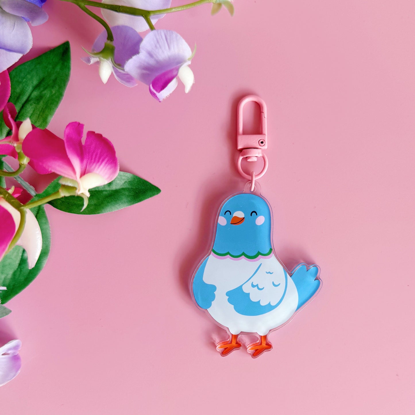 Blue cartoon pigeon keychain with a pink clip on a pink background with flowers.