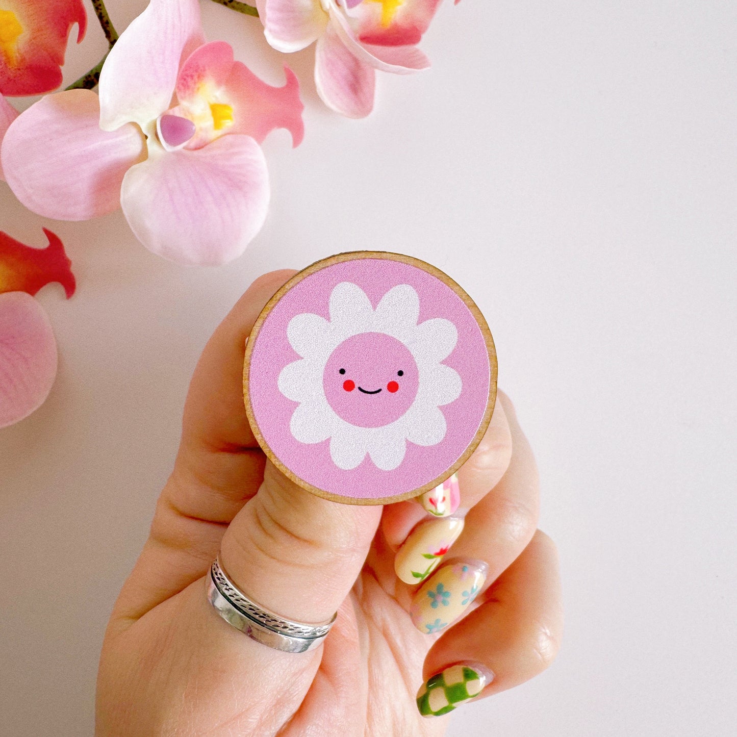 A pink pin badge, of a pink circle with a white daisy in the centre. the daisy is smiling and has rosy red cheeks. the backing card of the pin badge is yellow checkerboard patterned and has branding on.