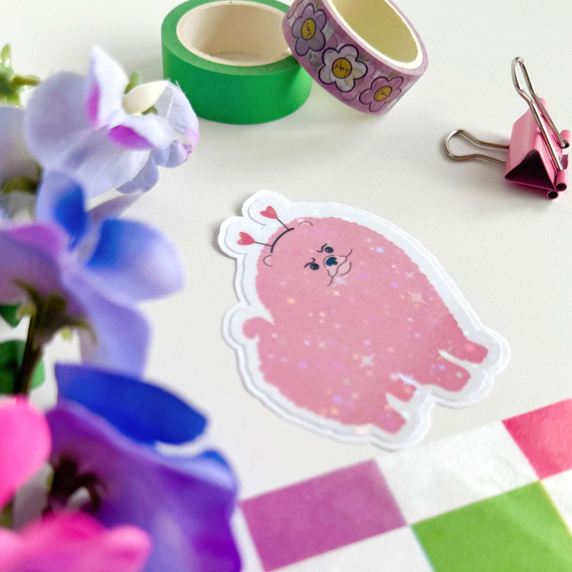A holographic sticker, of a pink grumpy looking dog with a heart shaped headband on. The sticker has an easy peel lip.