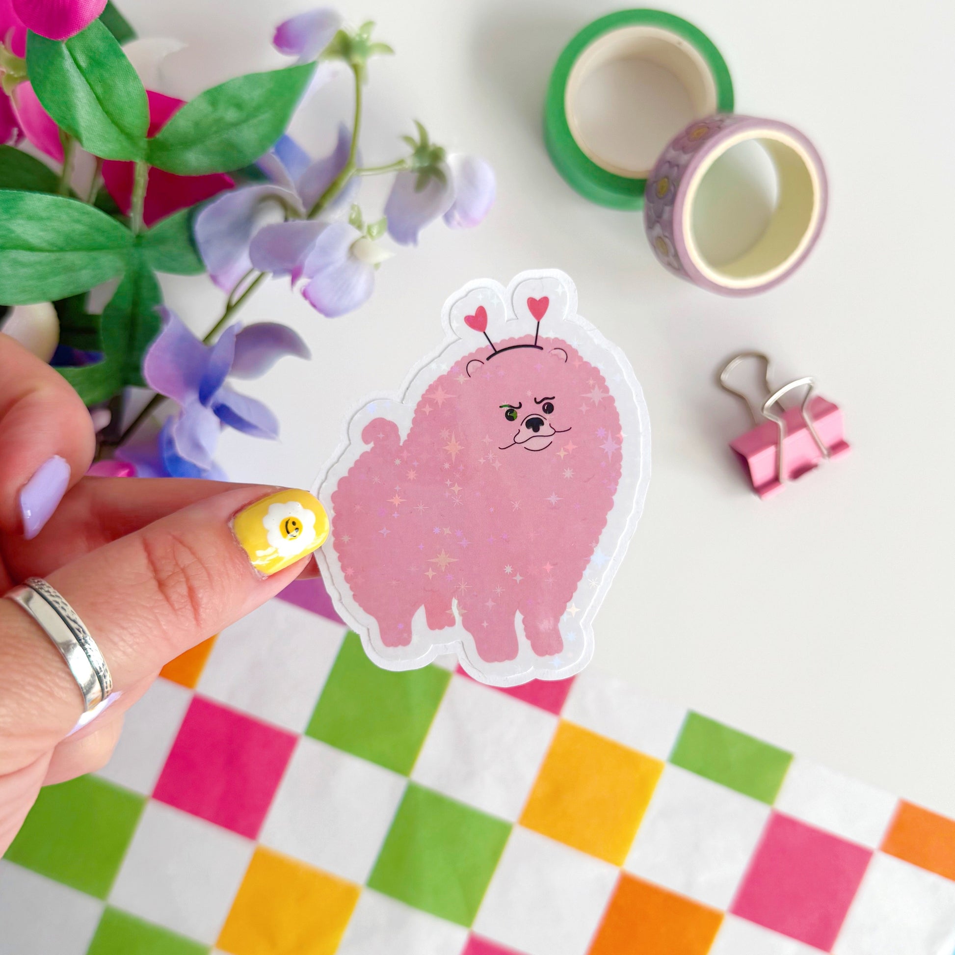 A holographic sticker, of a pink grumpy looking dog with a heart shaped headband on. The sticker has an easy peel lip.
