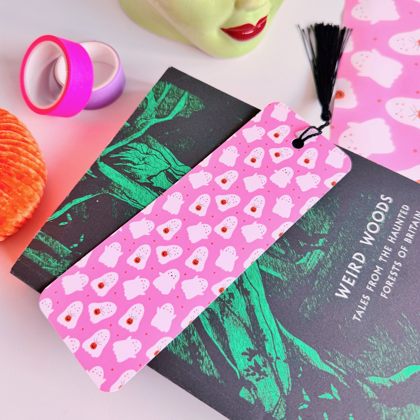 A styled flat-lay featuring a pink paper bookmark with a black tassel, printed with a repeating pattern of white ghosts with pumpkin trick or treat baskets.
