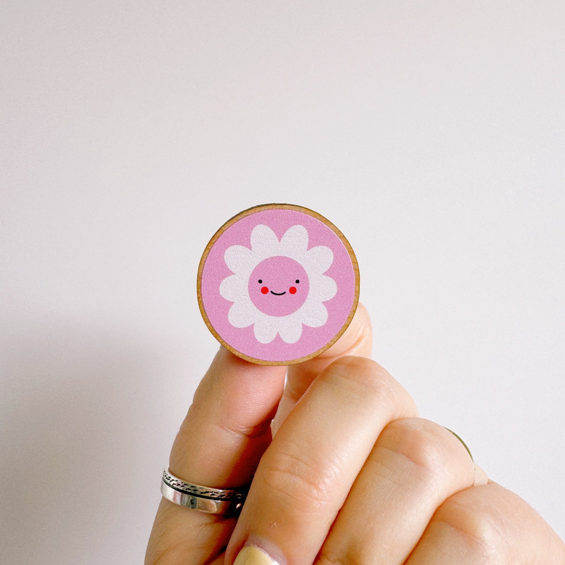 A pink pin badge, of a pink circle with a white daisy in the centre. the daisy is smiling and has rosy red cheeks. the backing card of the pin badge is yellow checkerboard patterned and has branding on.