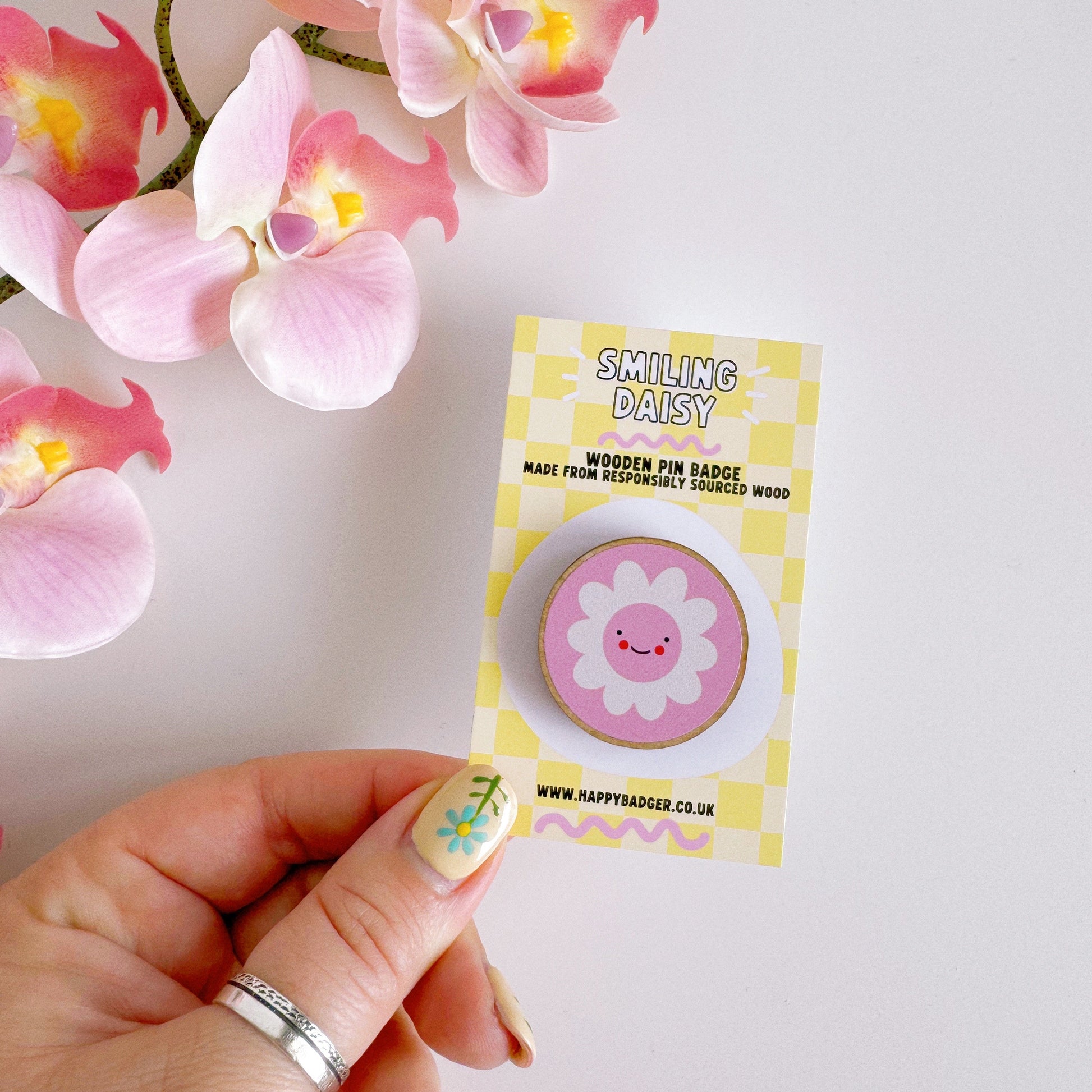 A pink pin badge, of a pink circle with a white daisy in the centre. the daisy is smiling and has rosy red cheeks. the backing card of the pin badge is yellow checkerboard patterned and has branding on.