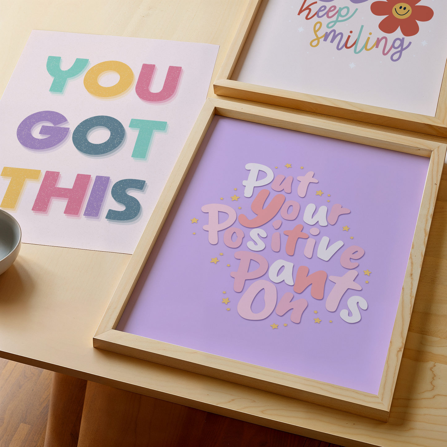 A purple quote print with text saying put your positive pants on, decorated with yellow stars. Shown with a You Got This Print and Keep Smiling Print in the background.