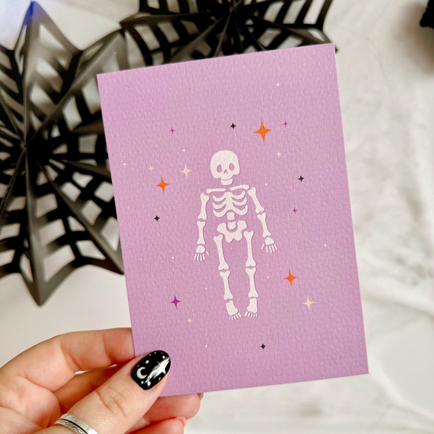 A halloween themed postcard of a cute spooky skeleton surrounded by stars, on a purple background.