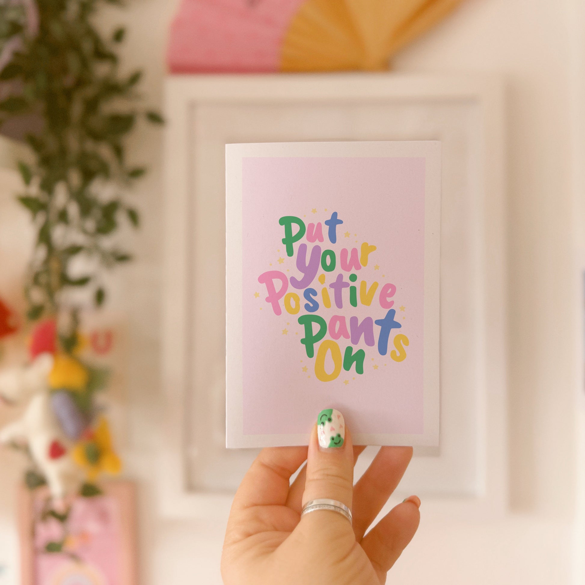 A colourful card with hand lettered text, saying 'put your positive pants on' on a light pink background.