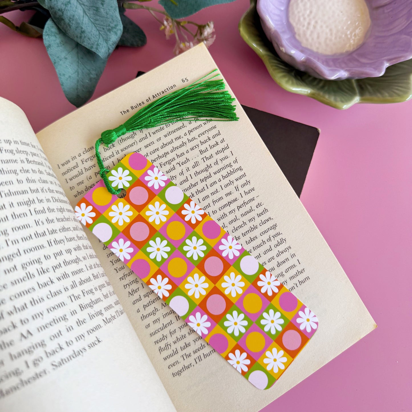 A brightly coloured retro look bookmark, featuring a checkerboard pattern with daisies and circles, in pink, green, yellow and orange. The bookmark has a coordinating green tassel.