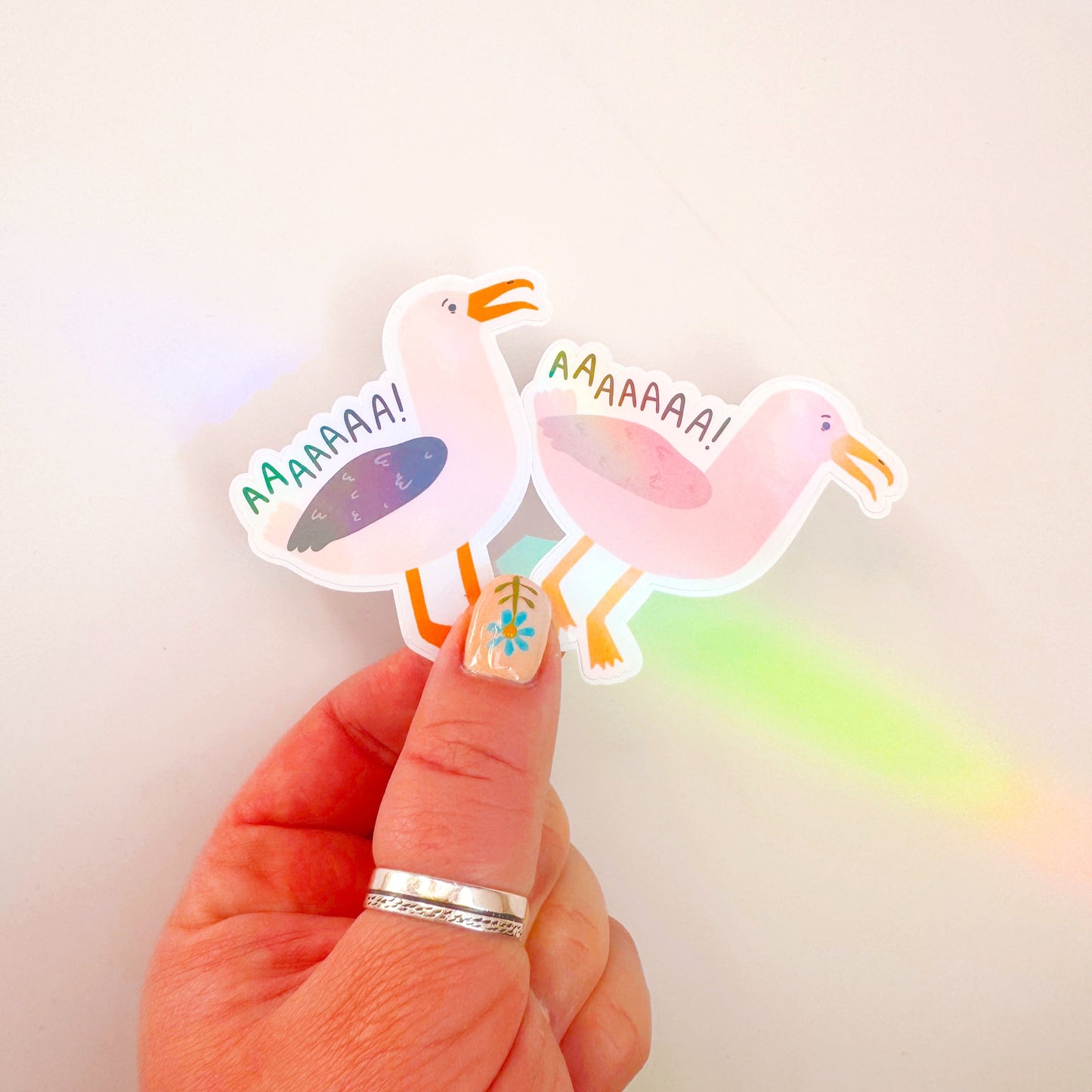 An image showing two holographic stickers of seagulls, one in pink one in more life-like colours. The text reads 'AAAAAAA!' as if the seagulls are screaming.