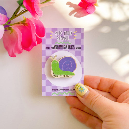 A cute wooden eco friendly pin badge, of a snail, that says 'taking it slow...'. The snail is purple and green and is smiling with rosy cheeks.