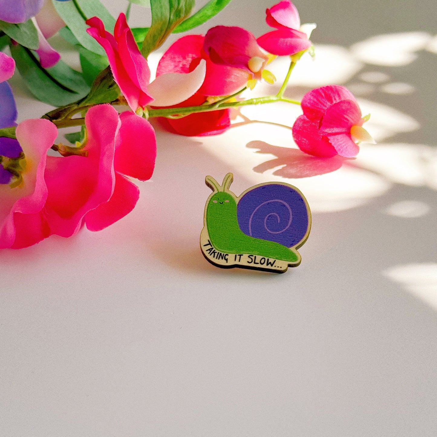 A cute wooden eco friendly pin badge, of a snail, that says 'taking it slow...'. The snail is purple and green and is smiling with rosy cheeks.