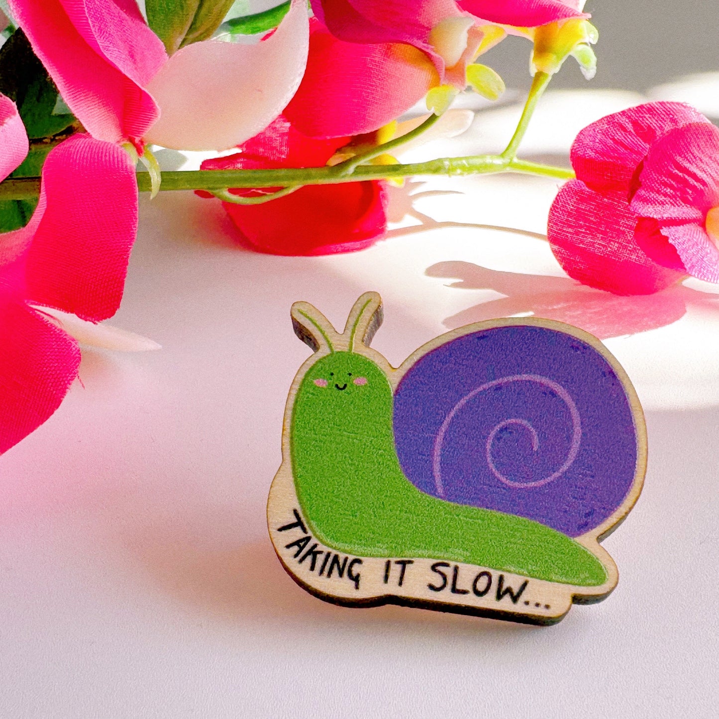 A cute wooden eco friendly pin badge, of a snail, that says 'taking it slow...'. The snail is purple and green and is smiling with rosy cheeks.