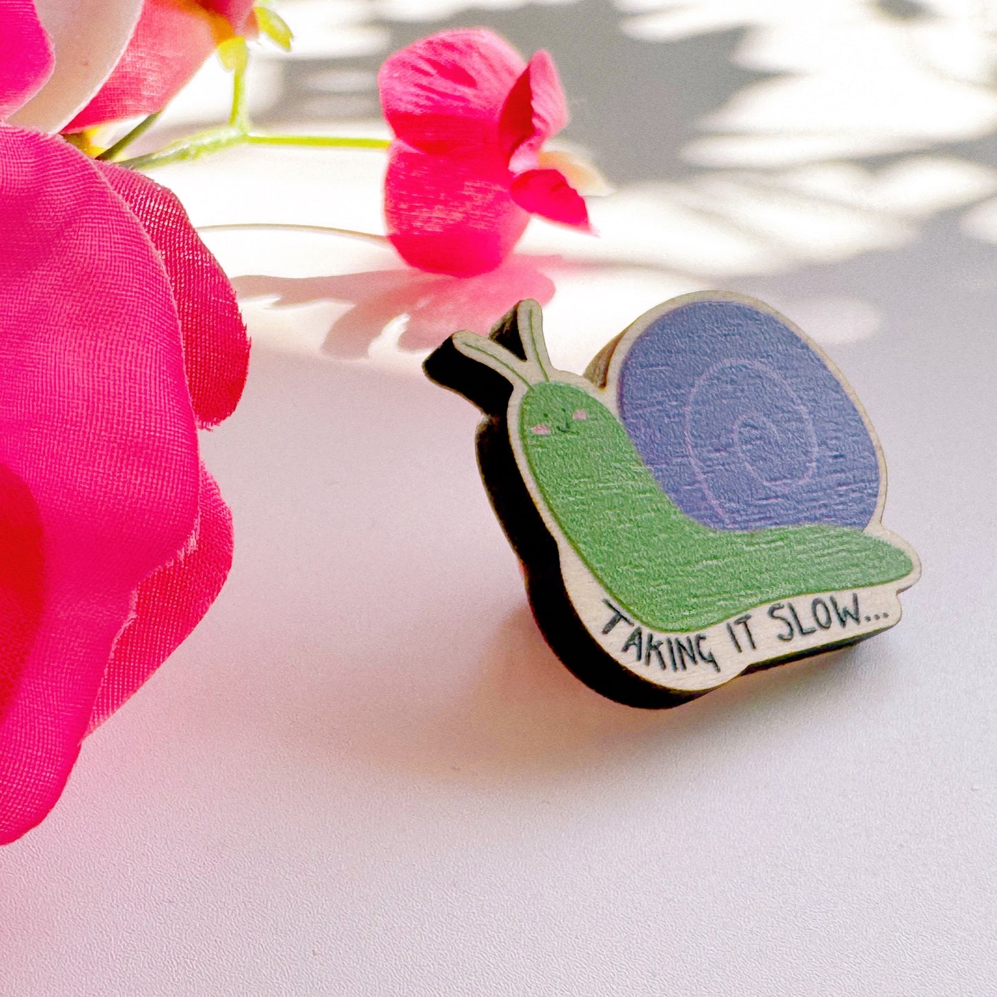 A cute wooden eco friendly pin badge, of a snail, that says 'taking it slow...'. The snail is purple and green and is smiling with rosy cheeks.
