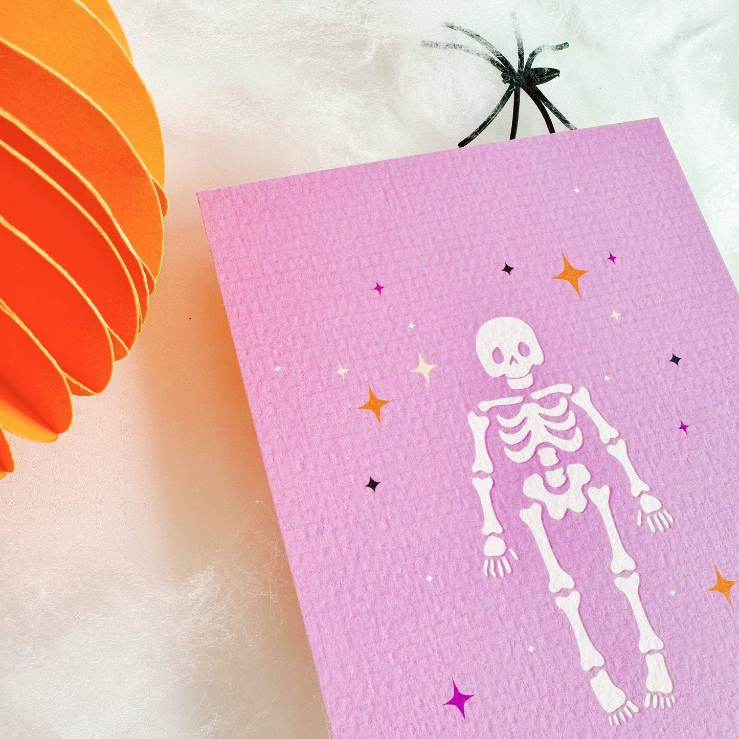 A halloween themed postcard of a cute spooky skeleton surrounded by stars, on a purple background.