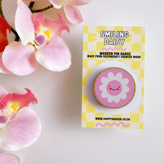 A pink pin badge, of a pink circle with a white daisy in the centre. the daisy is smiling and has rosy red cheeks. the backing card of the pin badge is yellow checkerboard patterned and has branding on.
