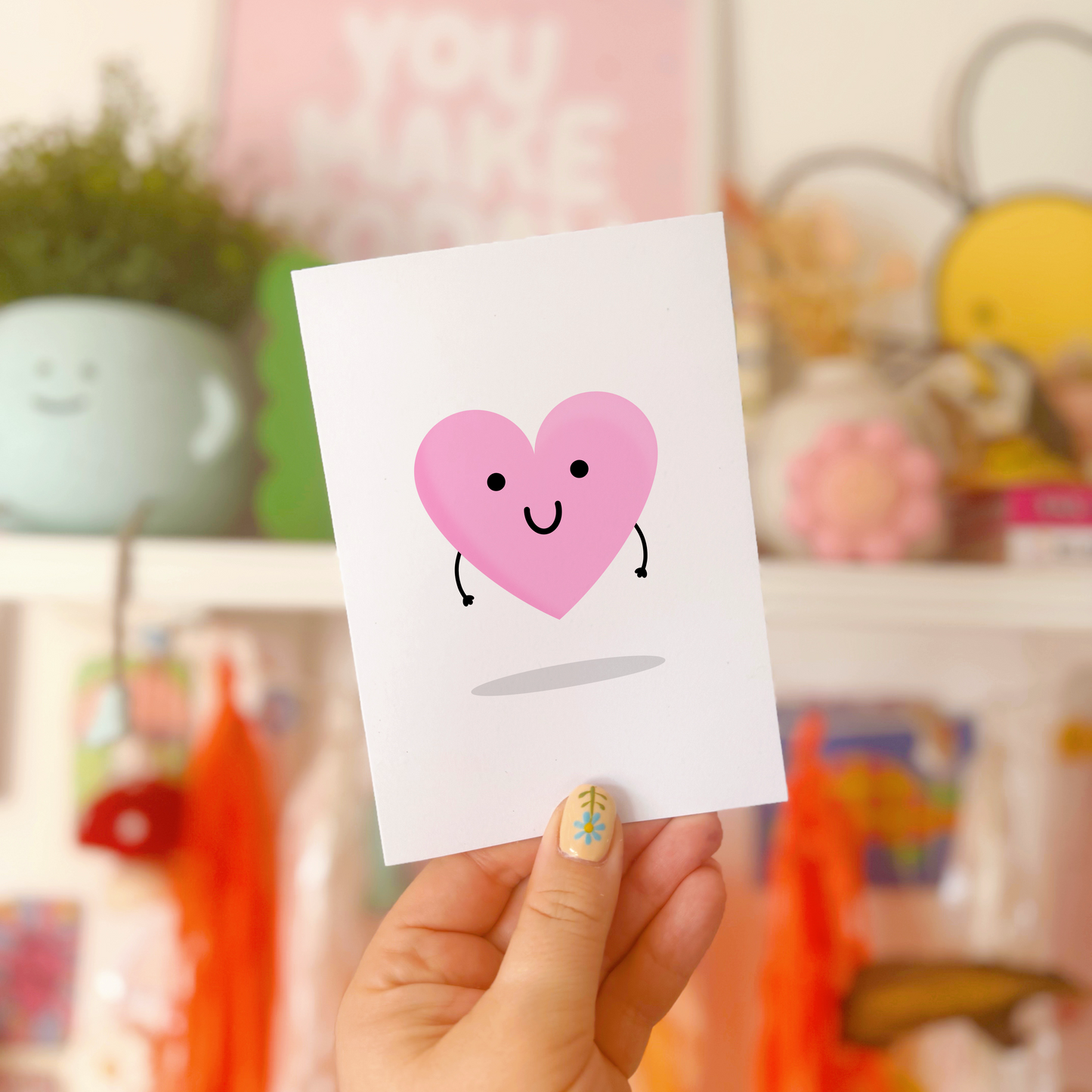 A pink love heart with arms and a smiling face, on a greetings card.