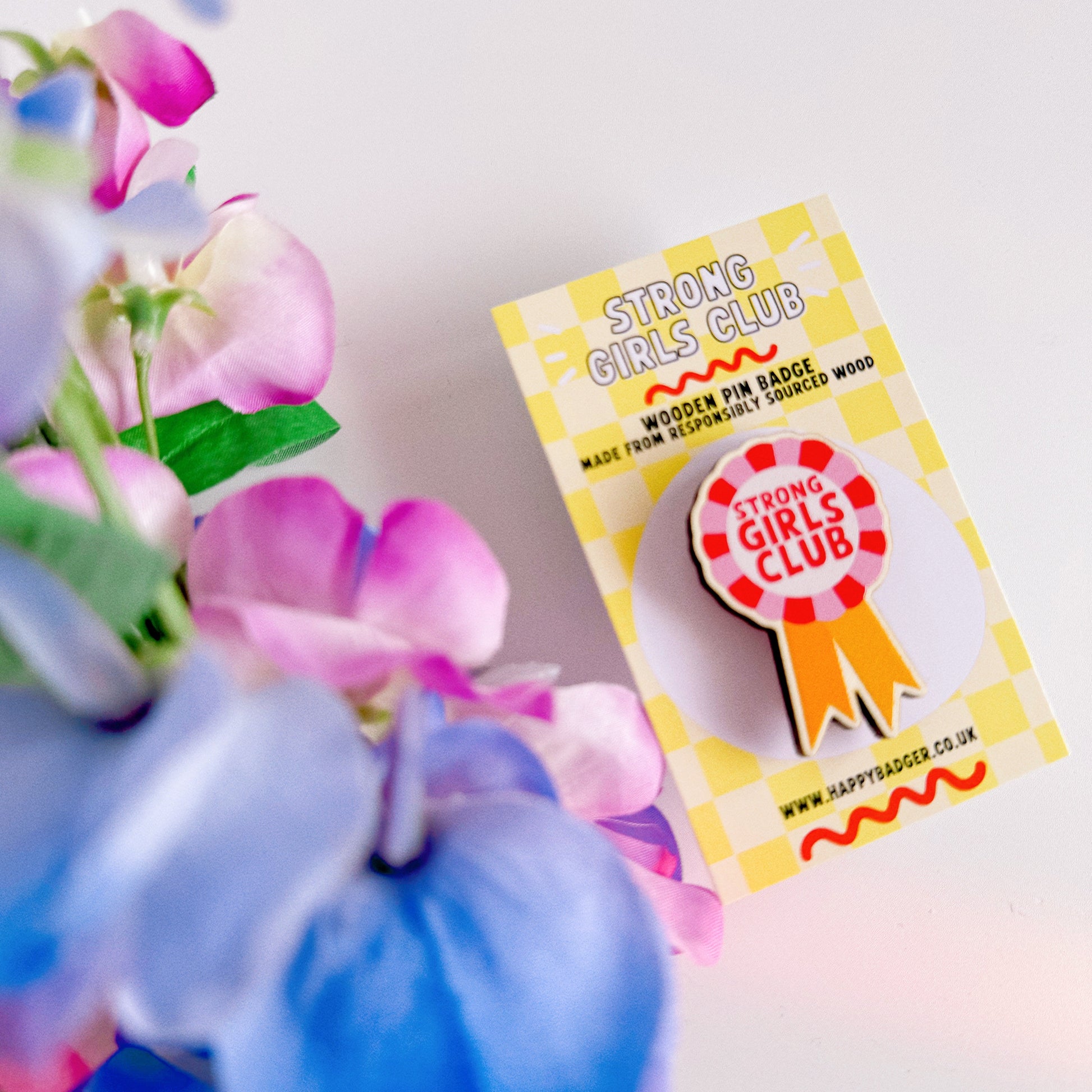 A wooden pin badge with a rosette in pink, red and yellow. The rosette has the words 'strong girls club' in the centre.
