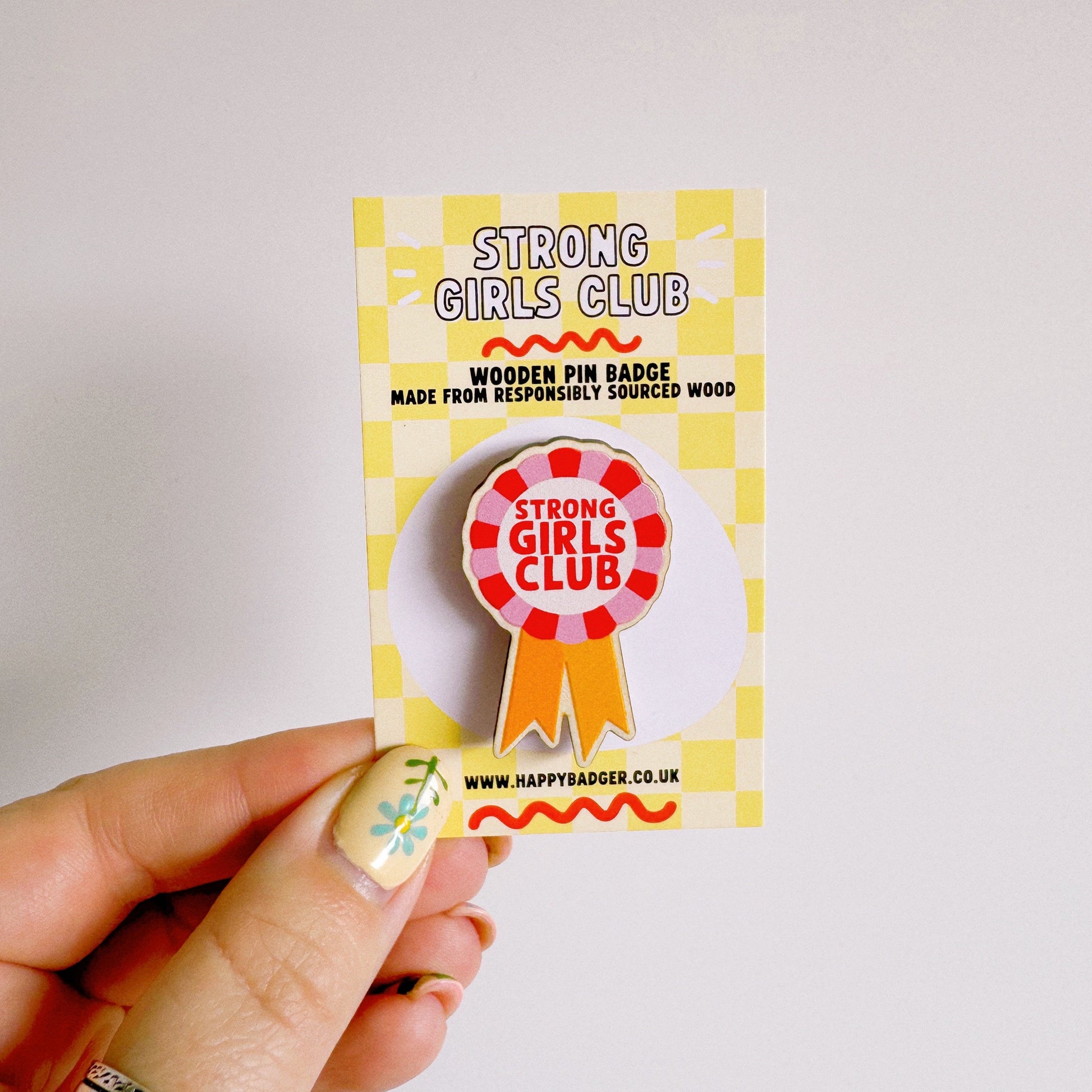 A wooden pin badge with a rosette in pink, red and yellow. The rosette has the words 'strong girls club' in the centre.
