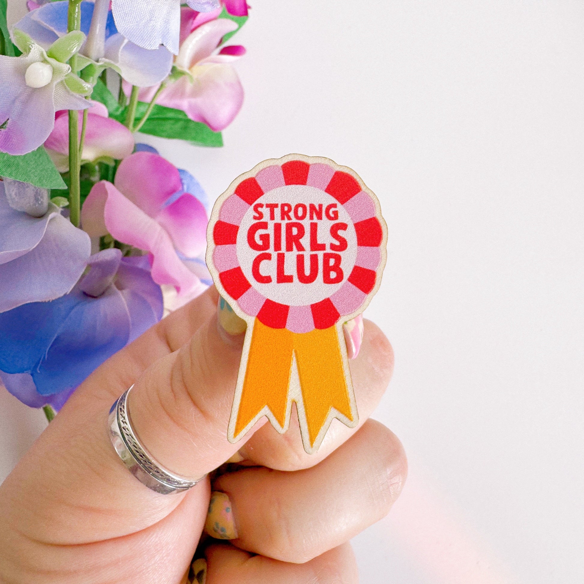 A wooden pin badge with a rosette in pink, red and yellow. The rosette has the words 'strong girls club' in the centre.