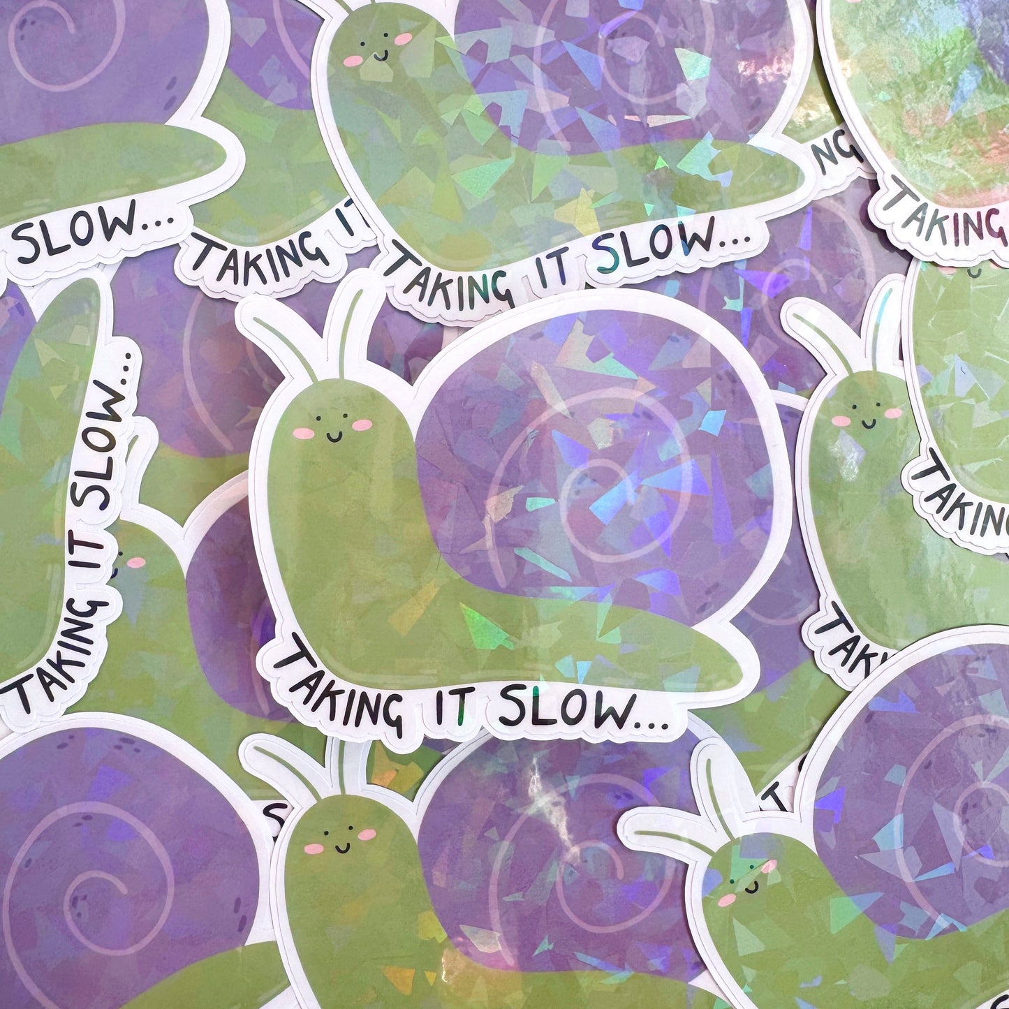 A holographic sticker of a purple and green smiling snail, with text underneath that reads 'taking it slow.'