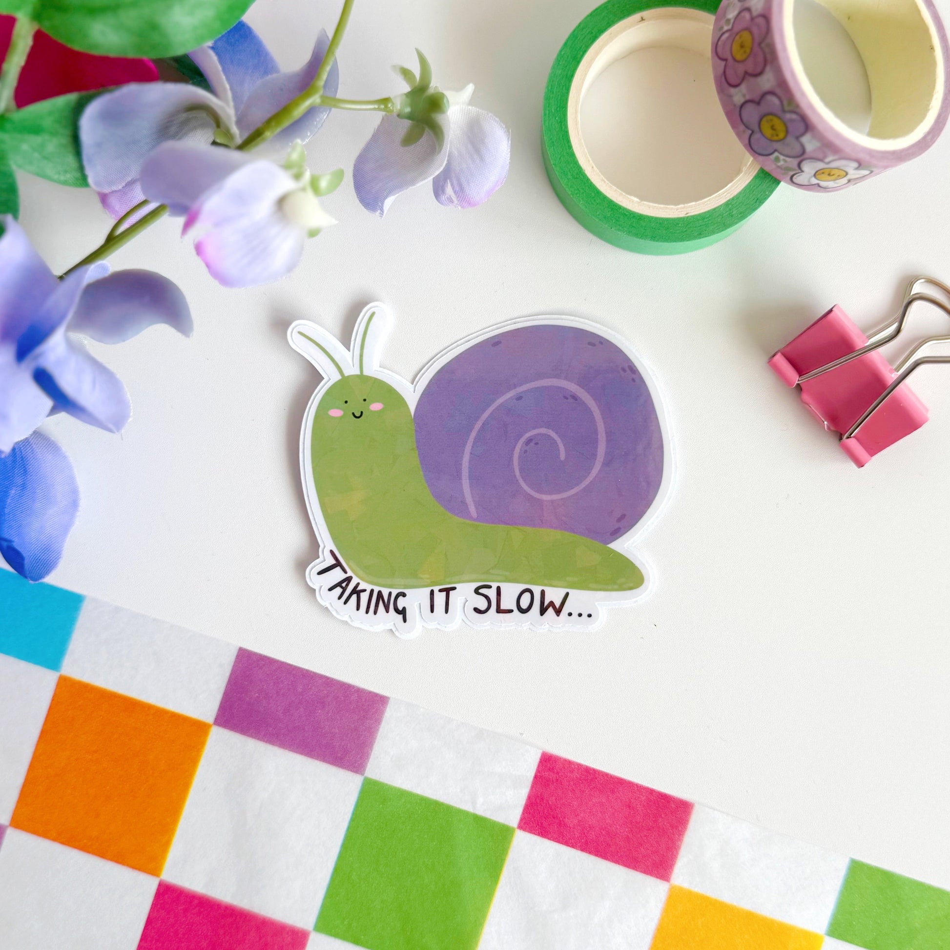 A holographic sticker of a purple and green smiling snail, with text underneath that reads 'taking it slow.'