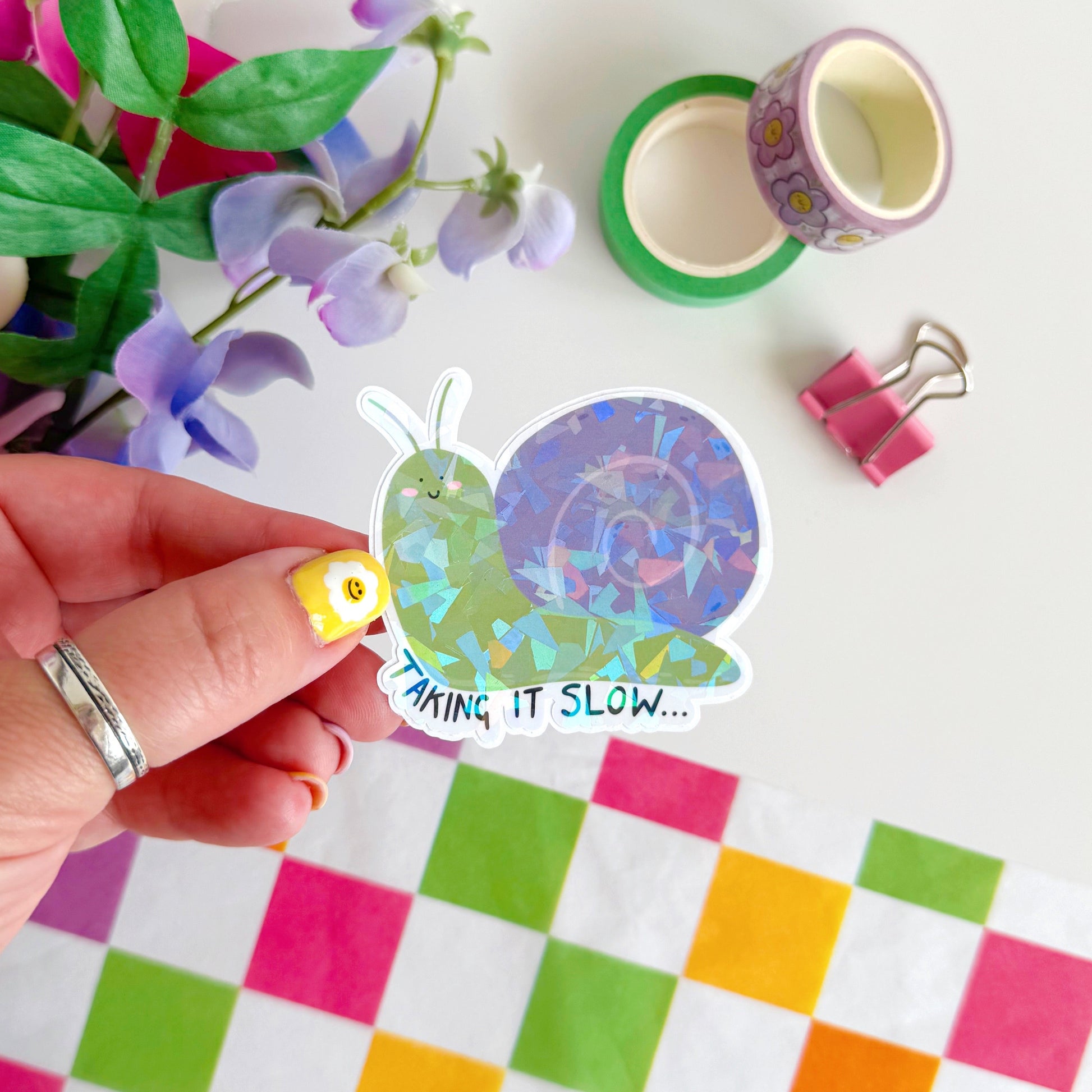 A holographic sticker of a purple and green smiling snail, with text underneath that reads 'taking it slow.'