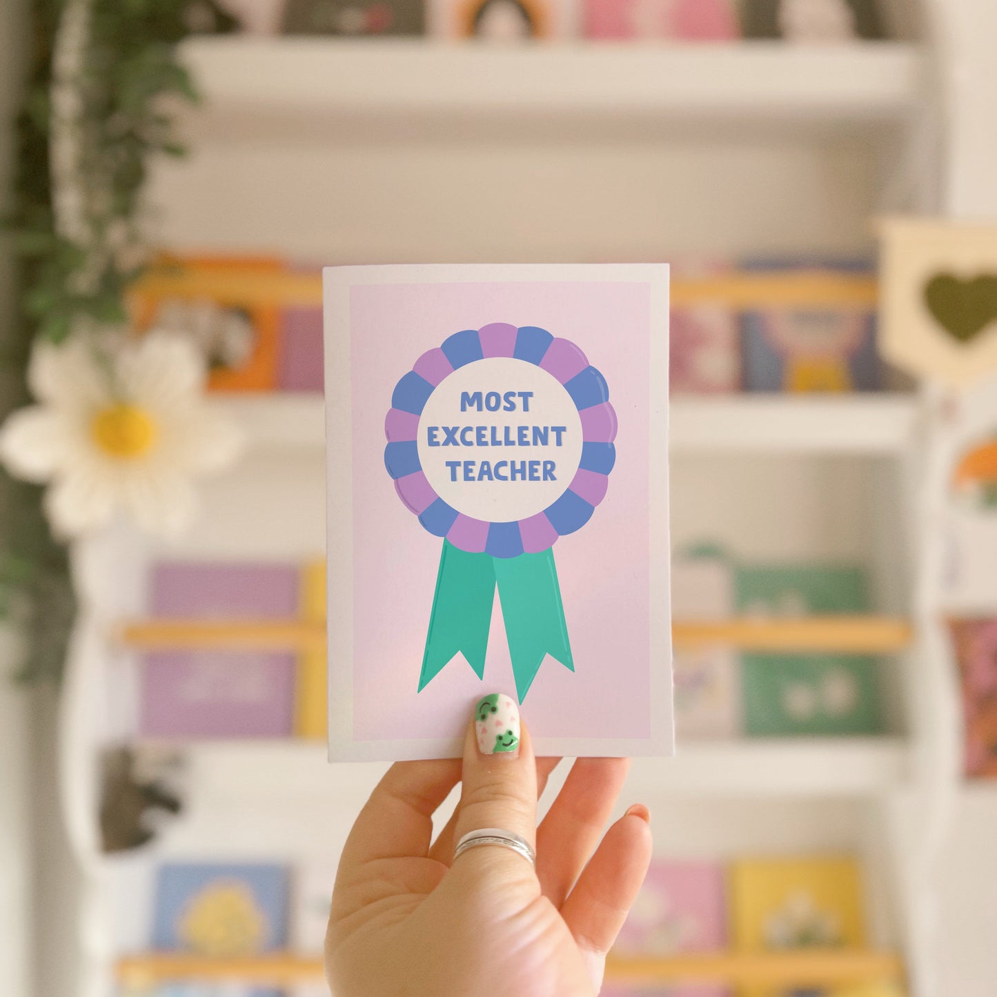 A greetings card with a rosette with text saying most excellent teacher. The colour combo is green blue and purple.