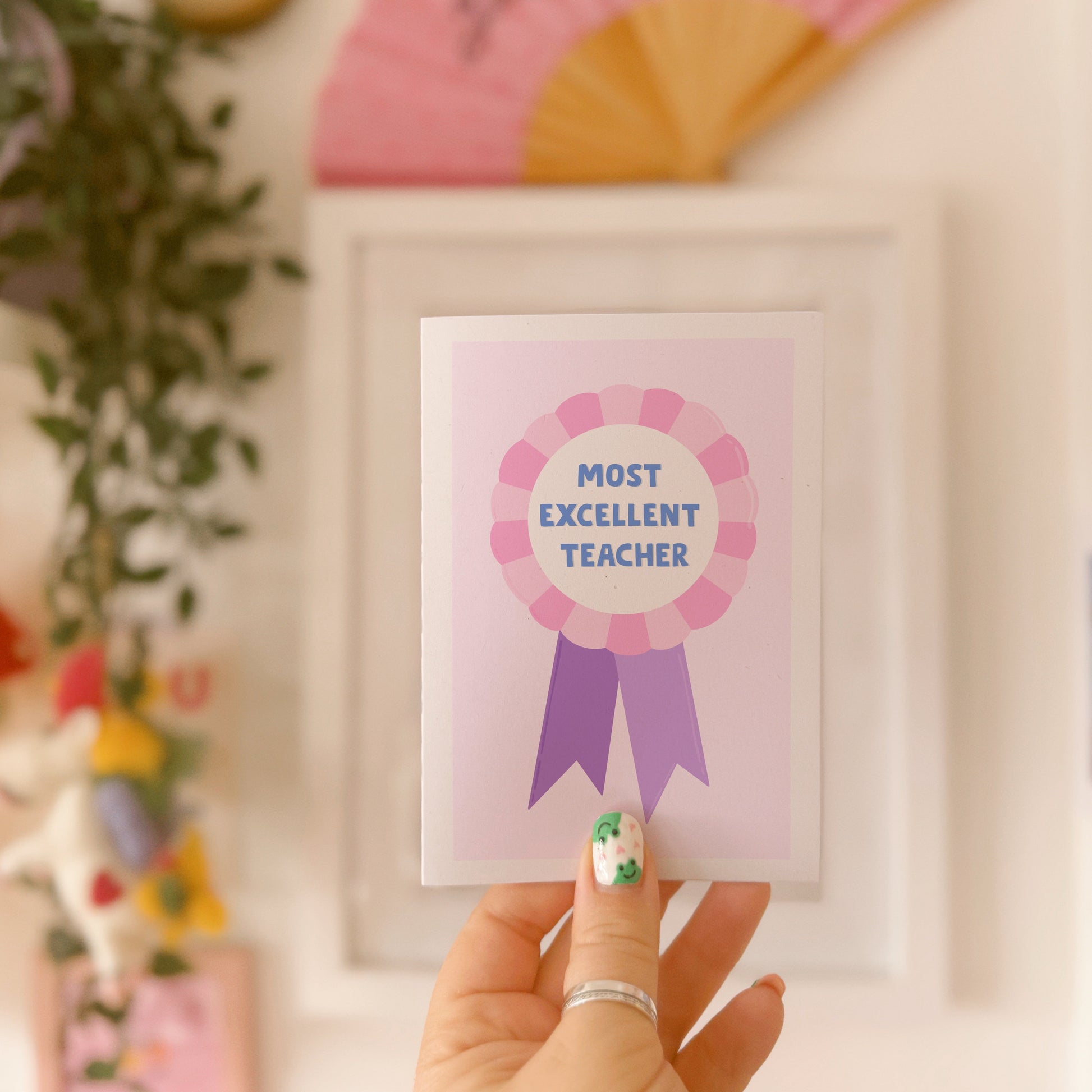 A greetings card with a rosette with text saying most excellent teacher. The colour combo is pink and purple