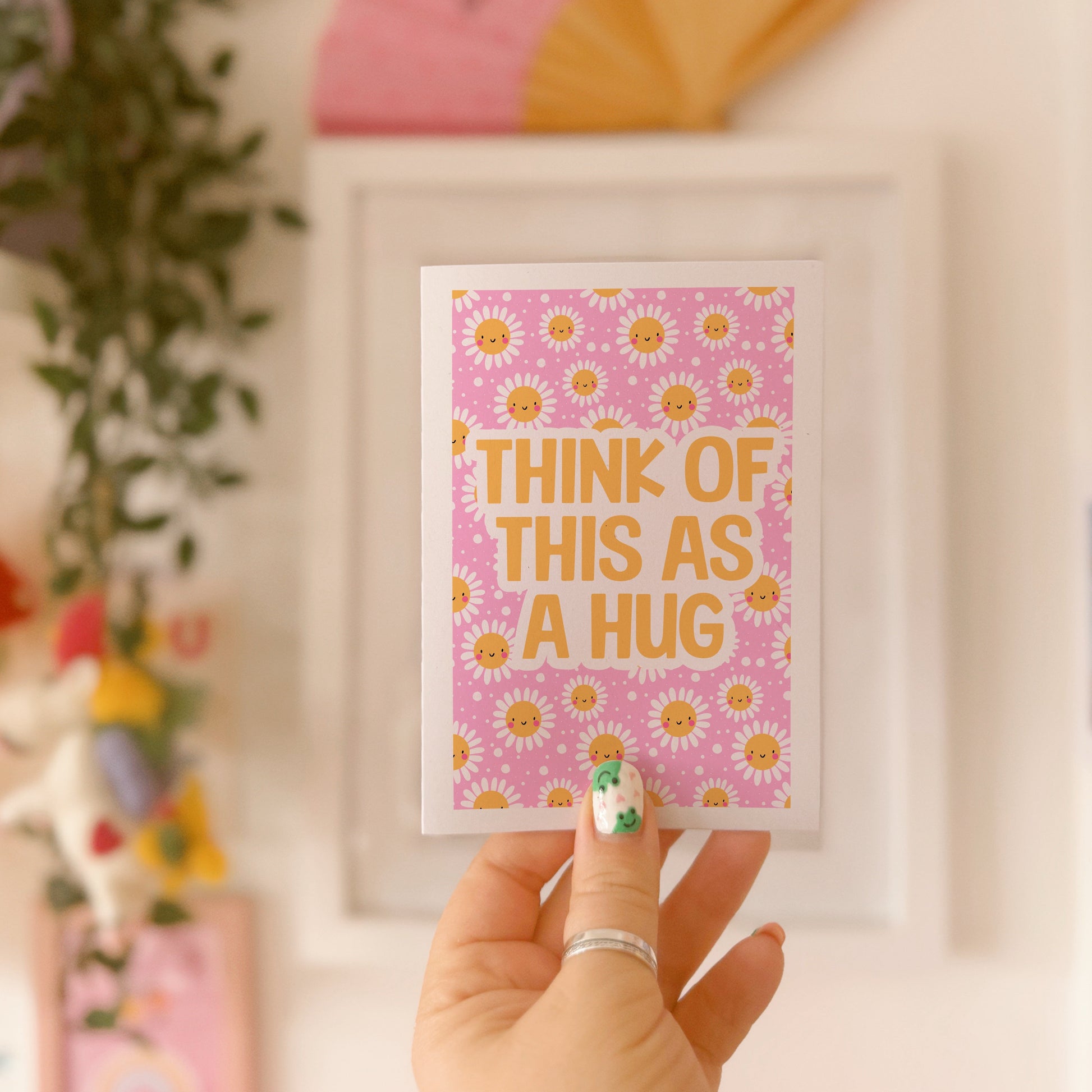 A bright pink and yellow greetings card, with a smiling daisy pattern. The text on the card reads 'think of this as a hug'.