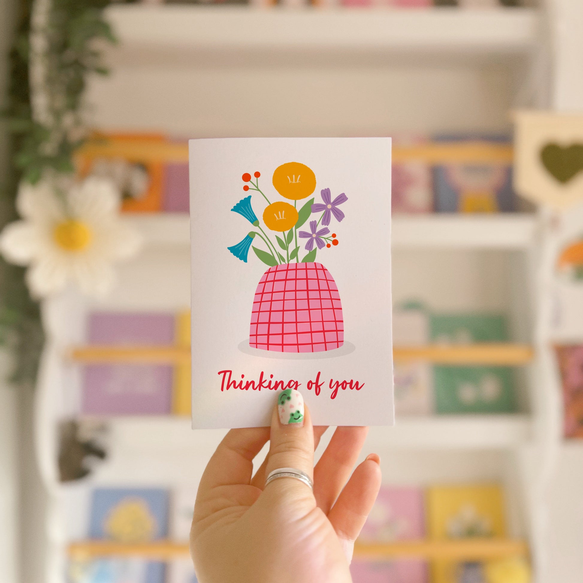 A card with a vase of colourful flowers, on a white background, with red hand written text underneath saying 'thinking of you'.