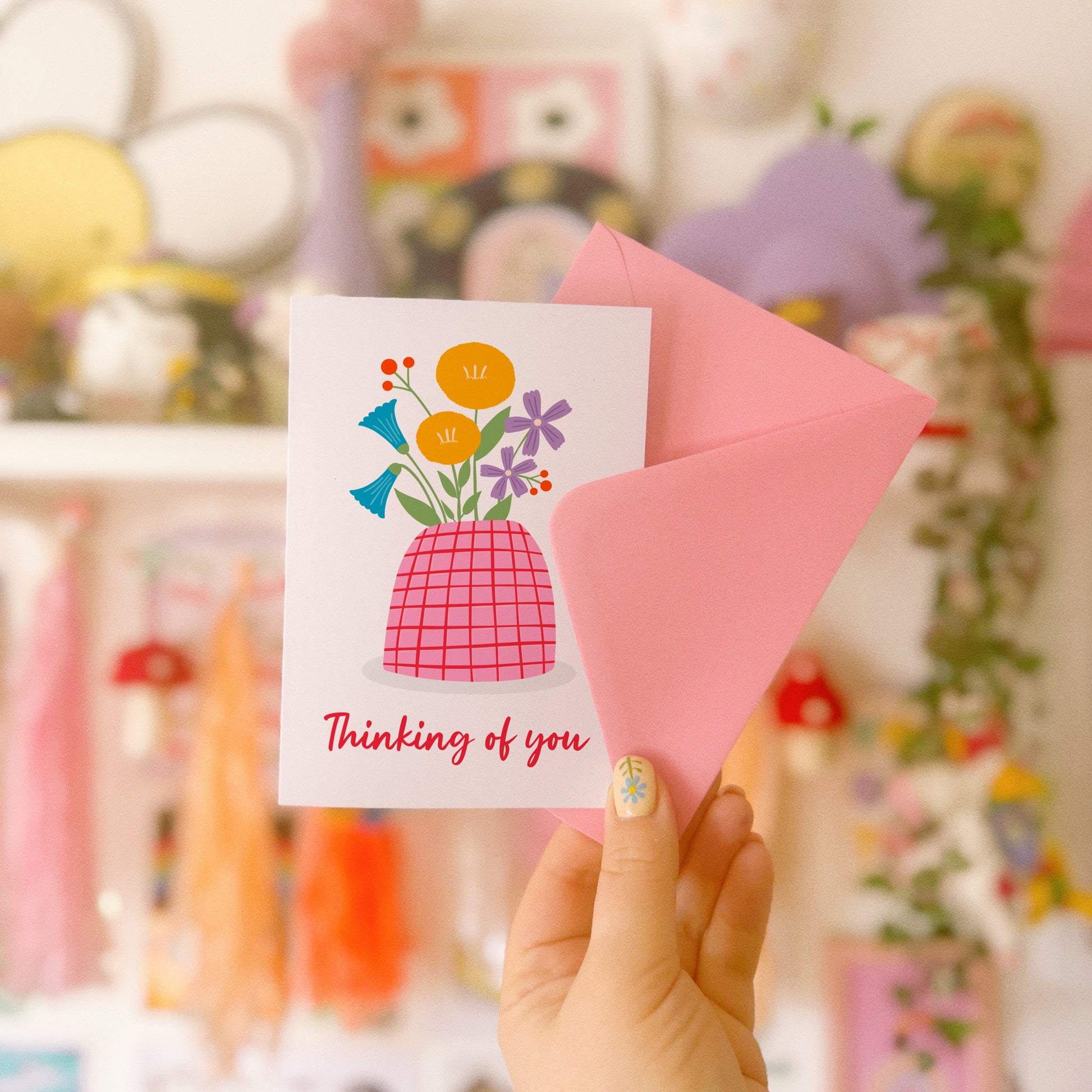 A card with a vase of colourful flowers, on a white background, with red hand written text underneath saying 'thinking of you'.