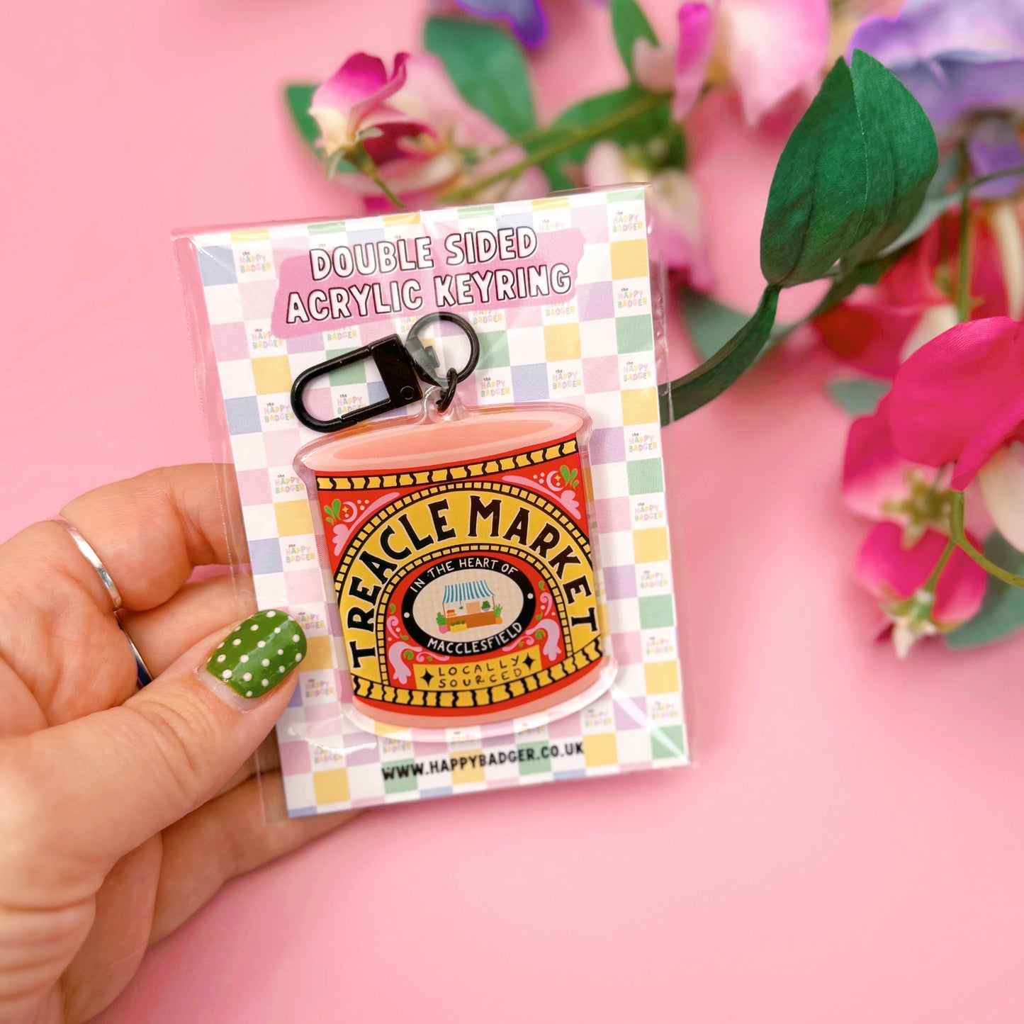 Hand holding a small Treacle Market tin keyring against a pink background with flowers.