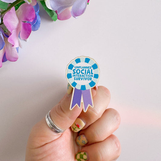 A wooden pin badge, shaped like an award rosette, that says 'unplanned social interaction survivor'.