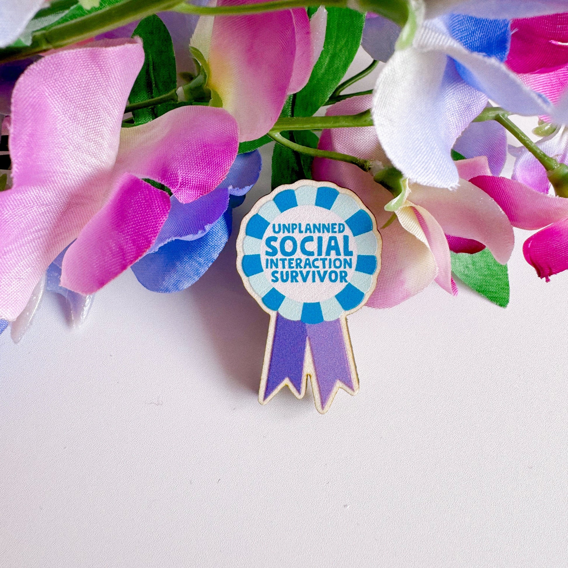 A wooden pin badge, shaped like an award rosette, that says 'unplanned social interaction survivor'.