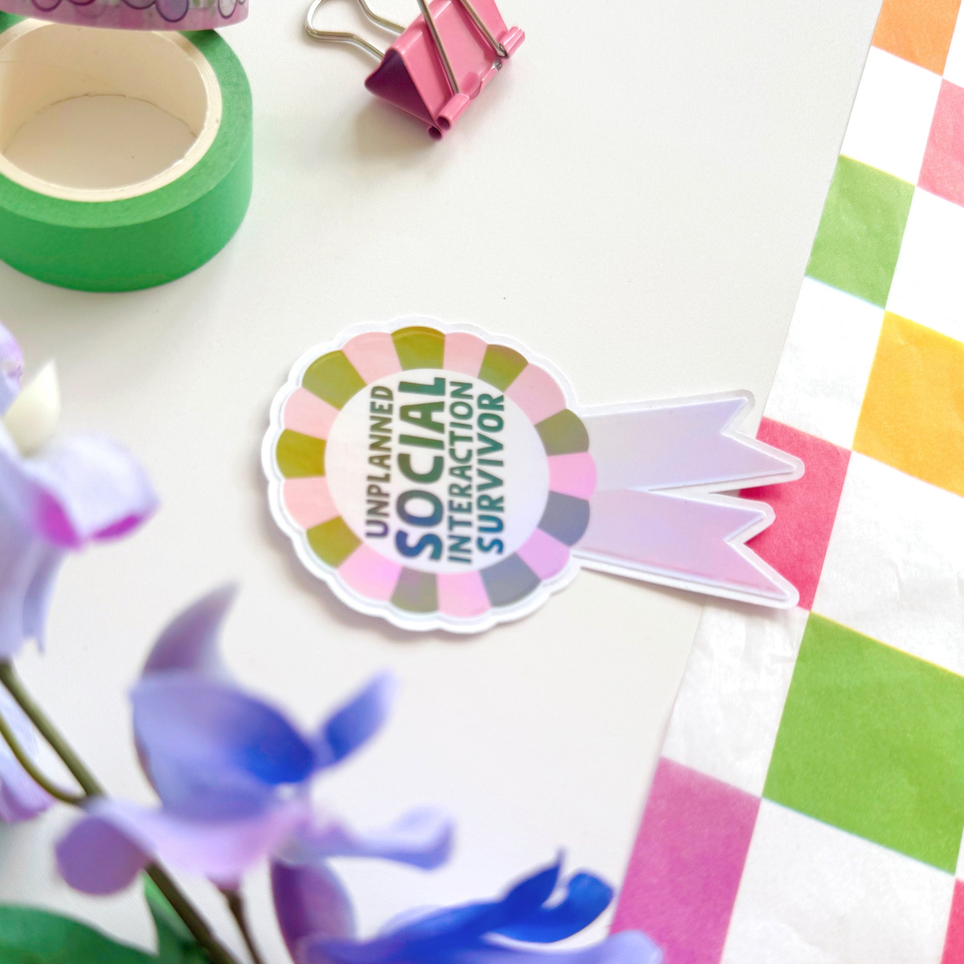 a sticker designed as a rosette, that says 'unplanned social interaction survivor' in pink and green.