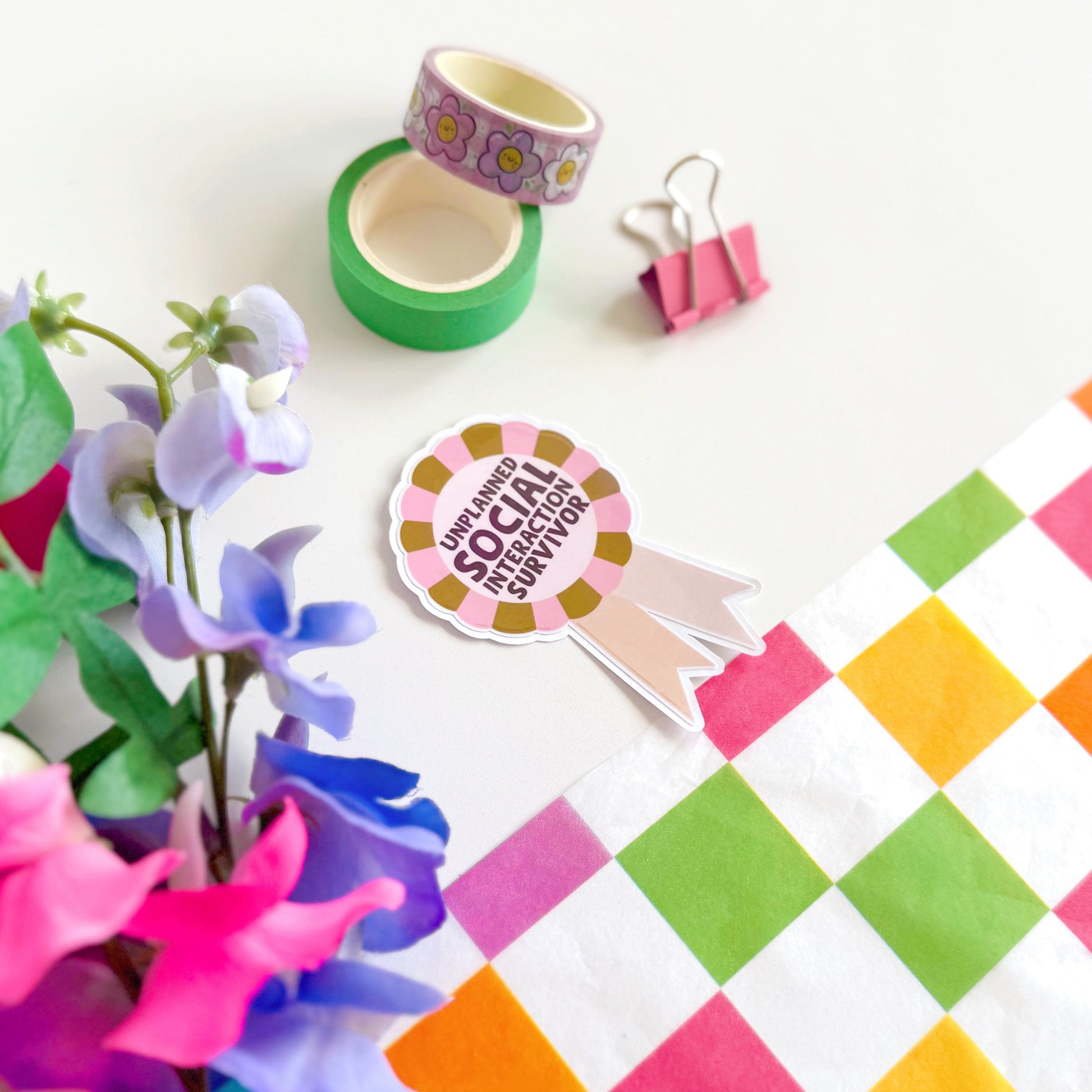 a sticker designed as a rosette, that says 'unplanned social interaction survivor' in pink and green.