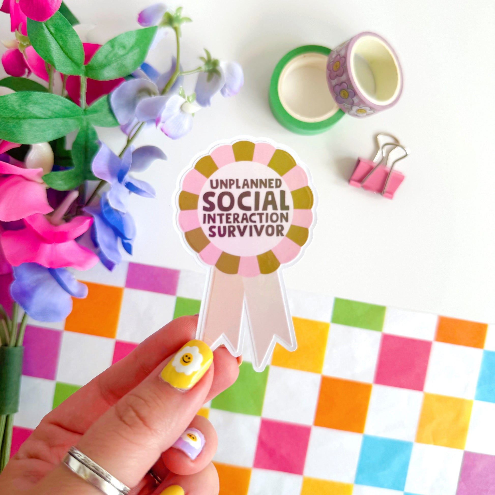 a sticker designed as a rosette, that says 'unplanned social interaction survivor' in pink and green.