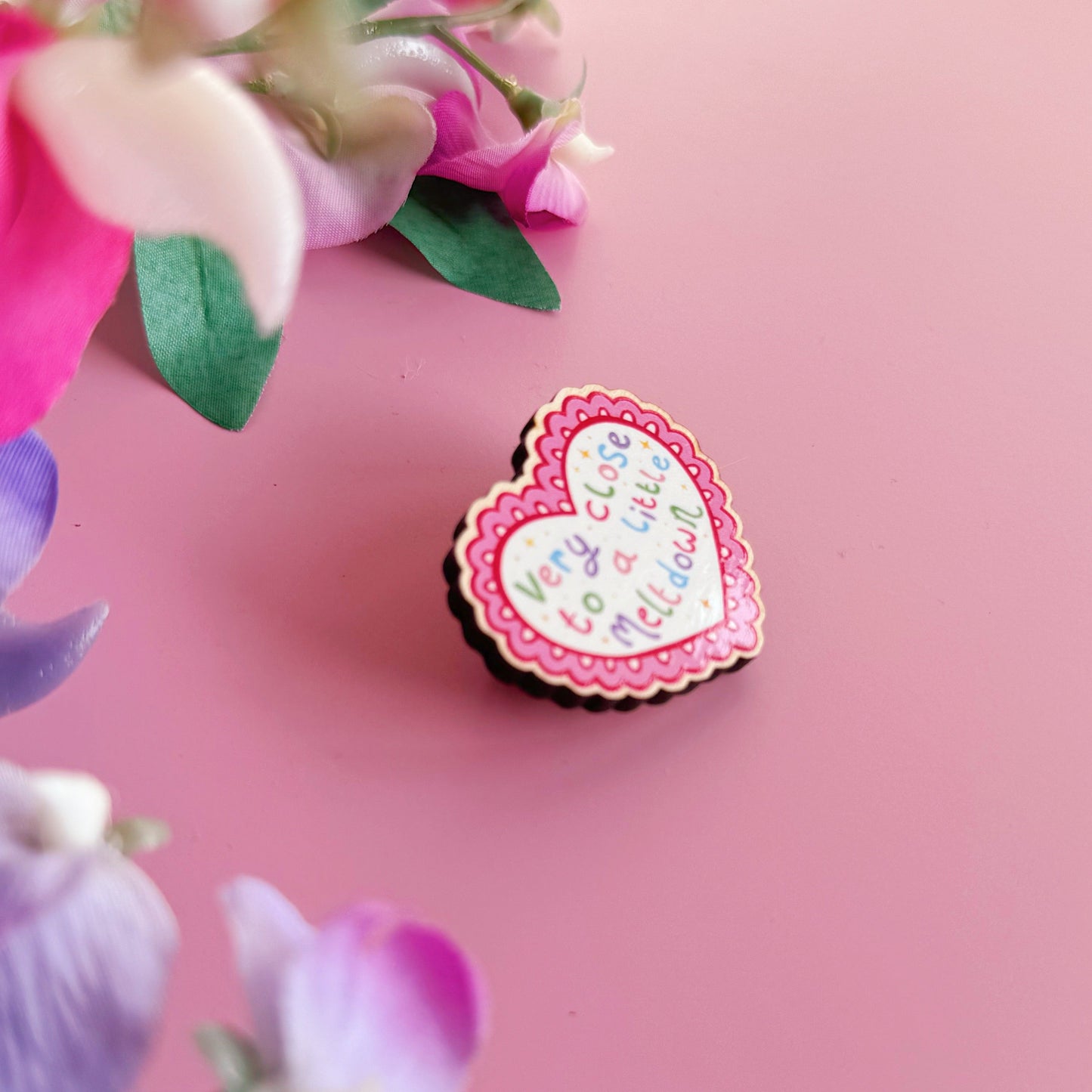 A wooden pin badge, with a glossy finish, in the shape of a heart with a funny quote in the centre.