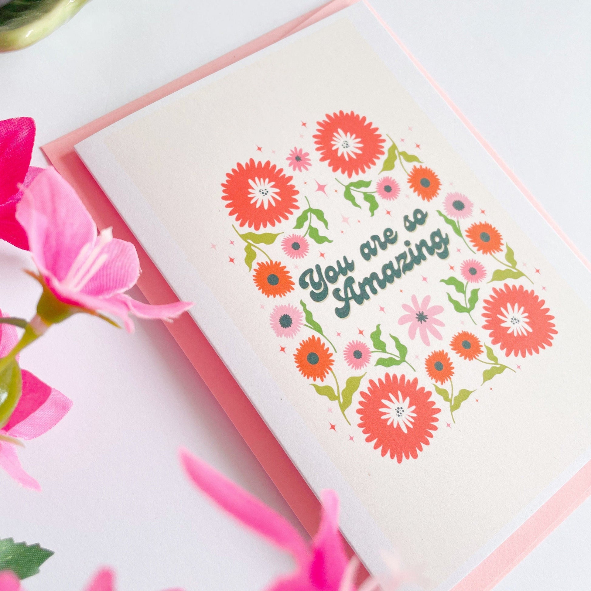 A greetings card with a cream background, with a symmetrical green orange and pink floral design. In the centre of the card are the words 'you are so amazing' in black text.