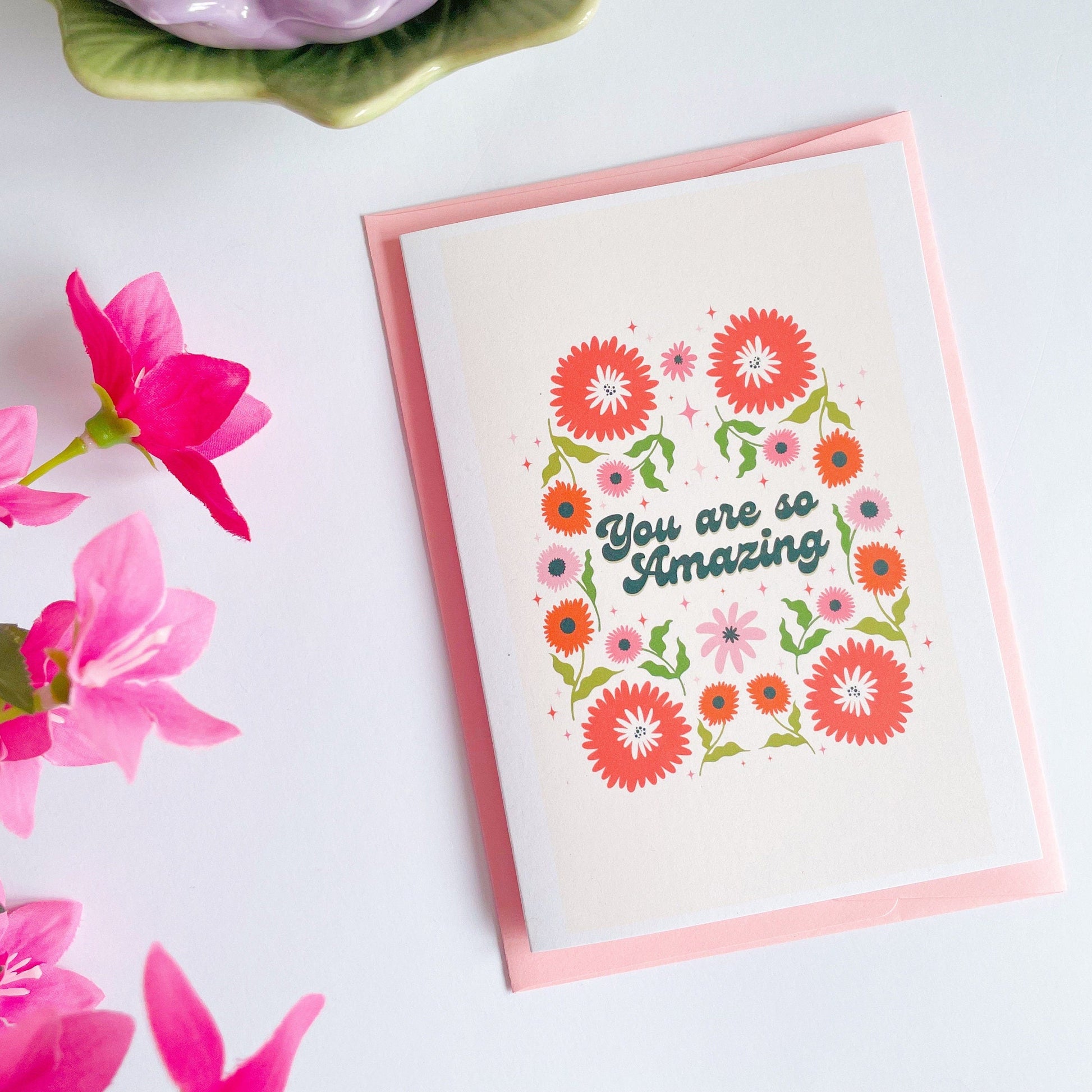 A greetings card with a cream background, with a symmetrical green orange and pink floral design. In the centre of the card are the words 'you are so amazing' in black text.