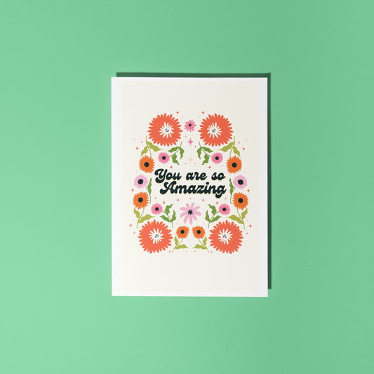 A greetings card with a cream background, with a symmetrical green orange and pink floral design. In the centre of the card are the words 'you are so amazing' in black text.