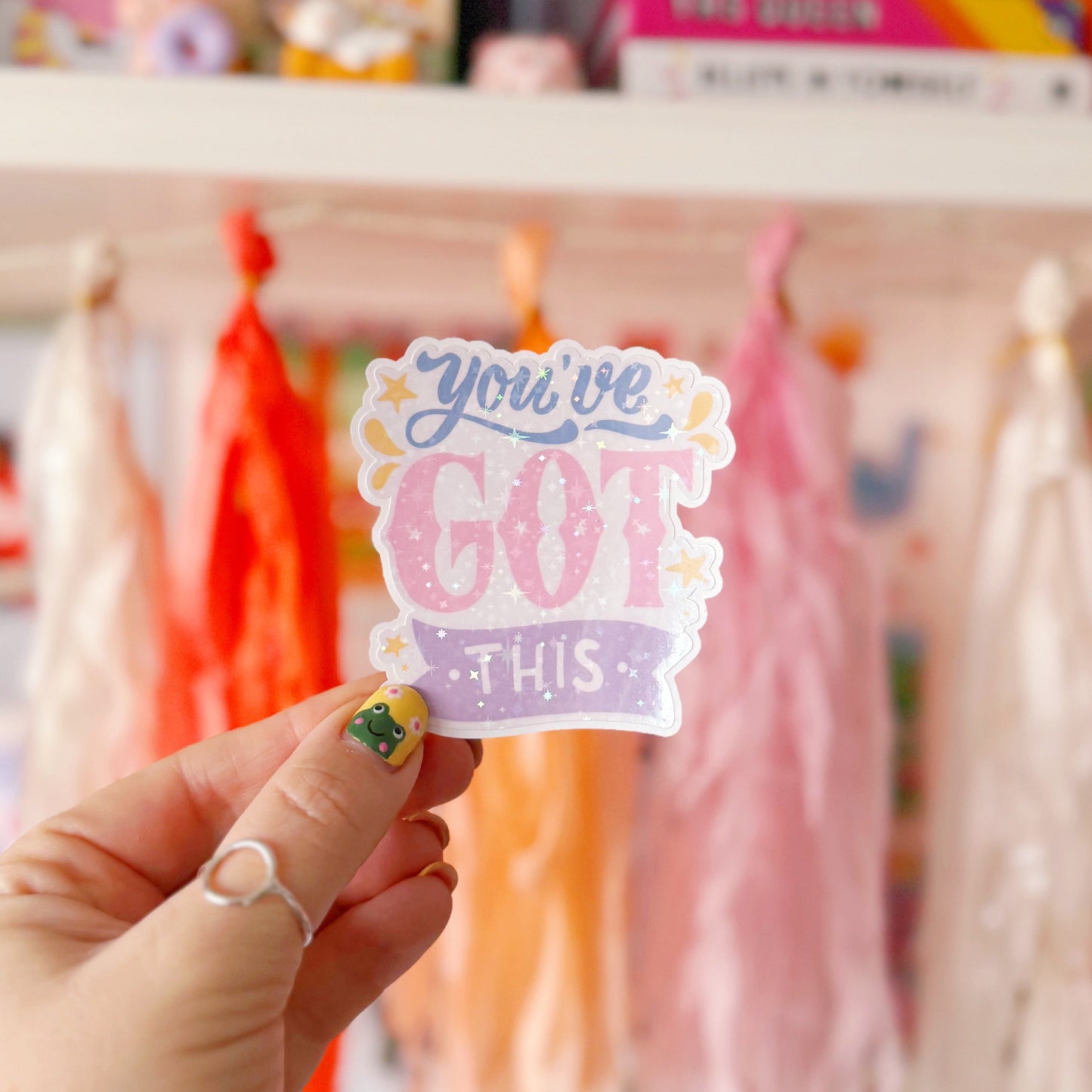A holographic sticker with script writing saying 'you've got this' in blue, pink and purple, with little yellow illustrated detailing around the text.
