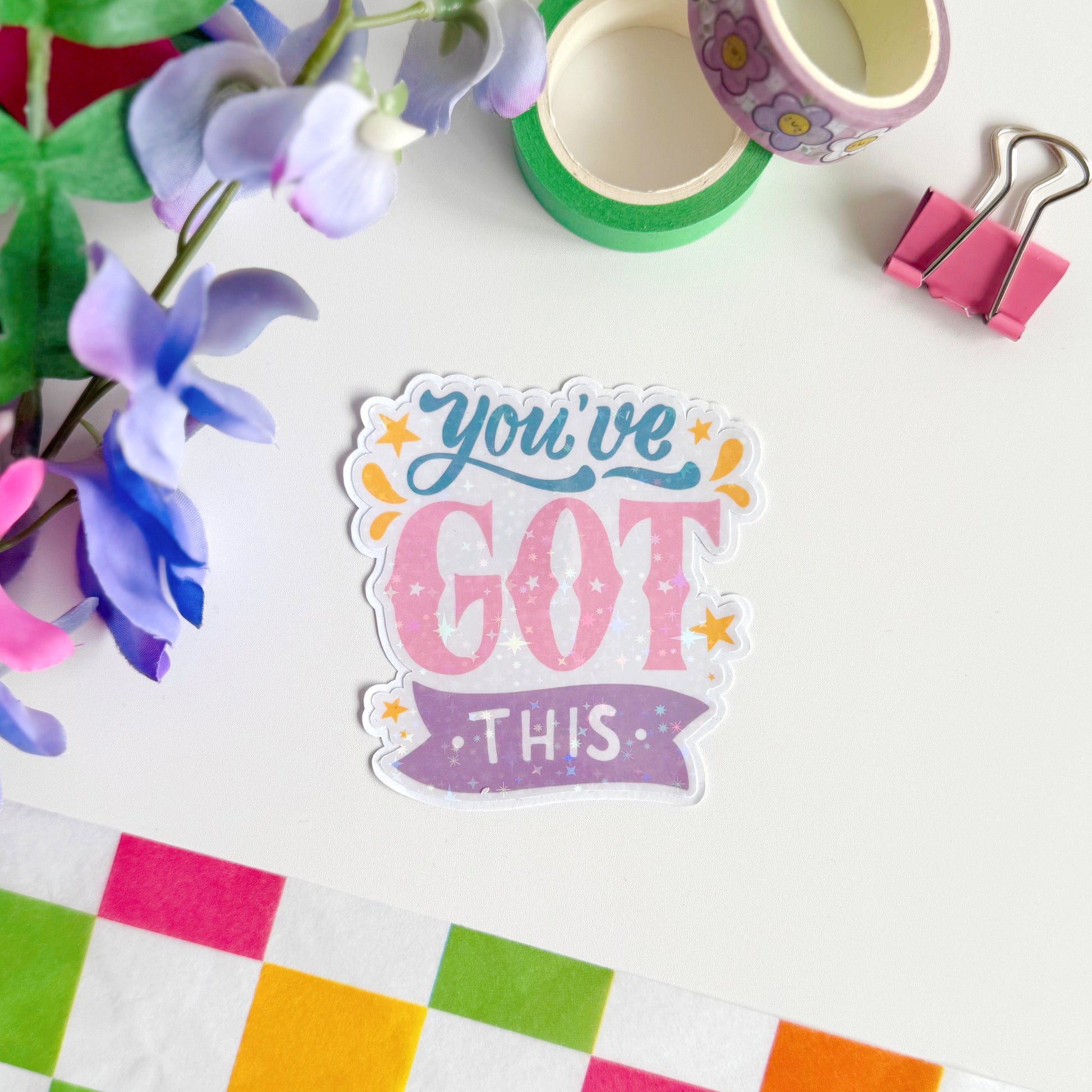 A holographic sticker with script writing saying 'you've got this' in blue, pink and purple, with little yellow illustrated detailing around the text.