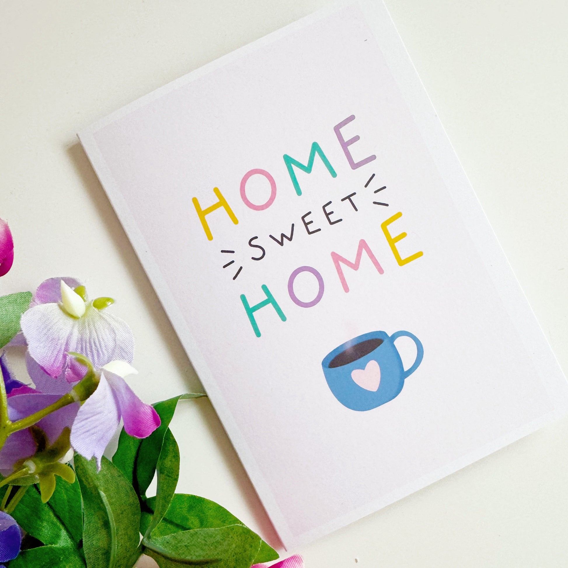 a pink card with text reading 'home sweet home' and a blue mug underneath with a heart on it.
