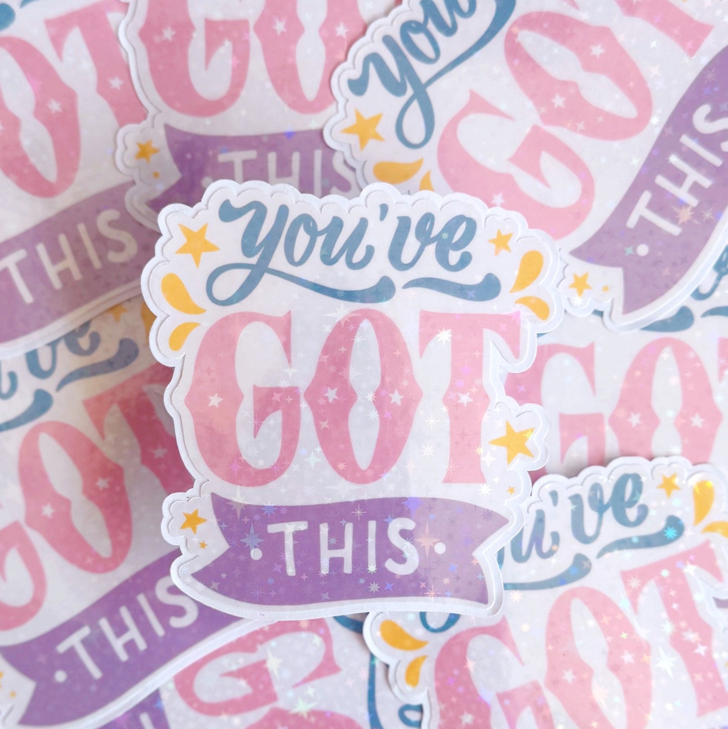 A holographic sticker with script writing saying 'you've got this' in blue, pink and purple, with little yellow illustrated detailing around the text.