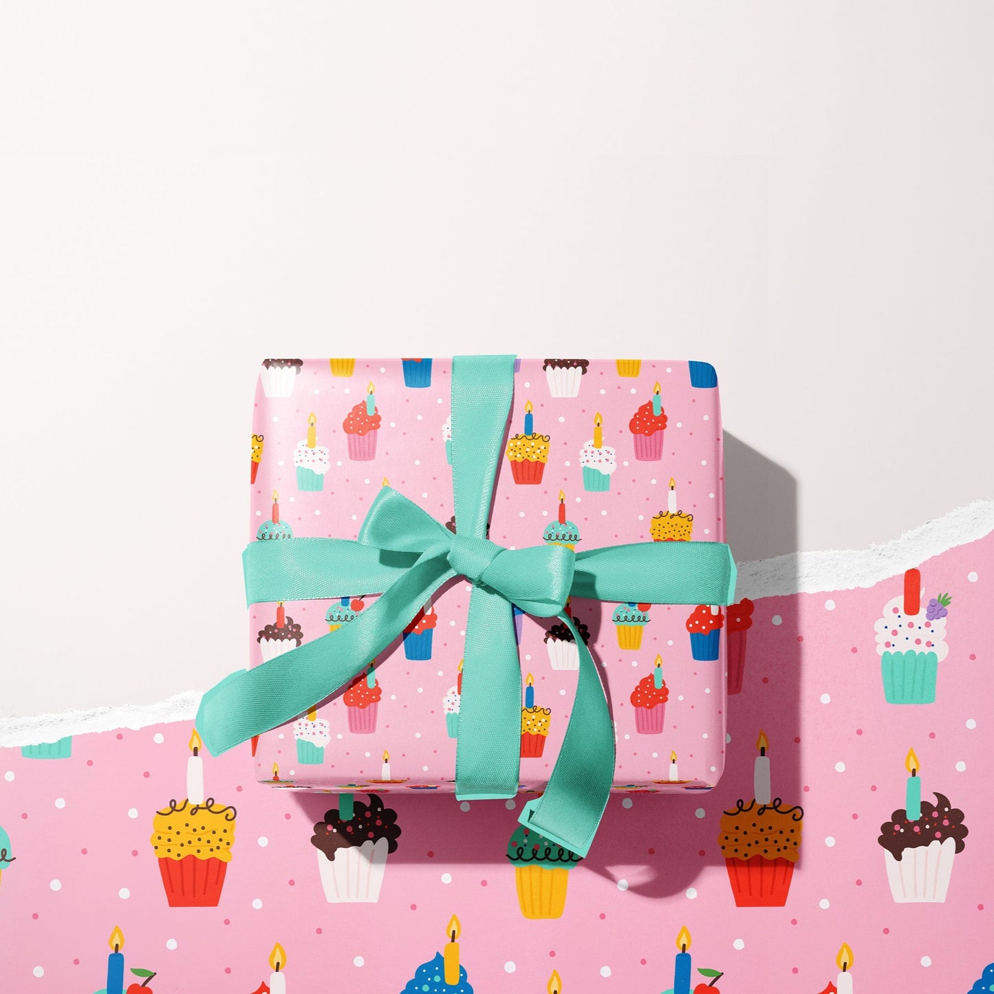 A present wrapped in pink gift wrap, which has colourful cupcakes and candles all over. The present is wrapped with a teal bow.
