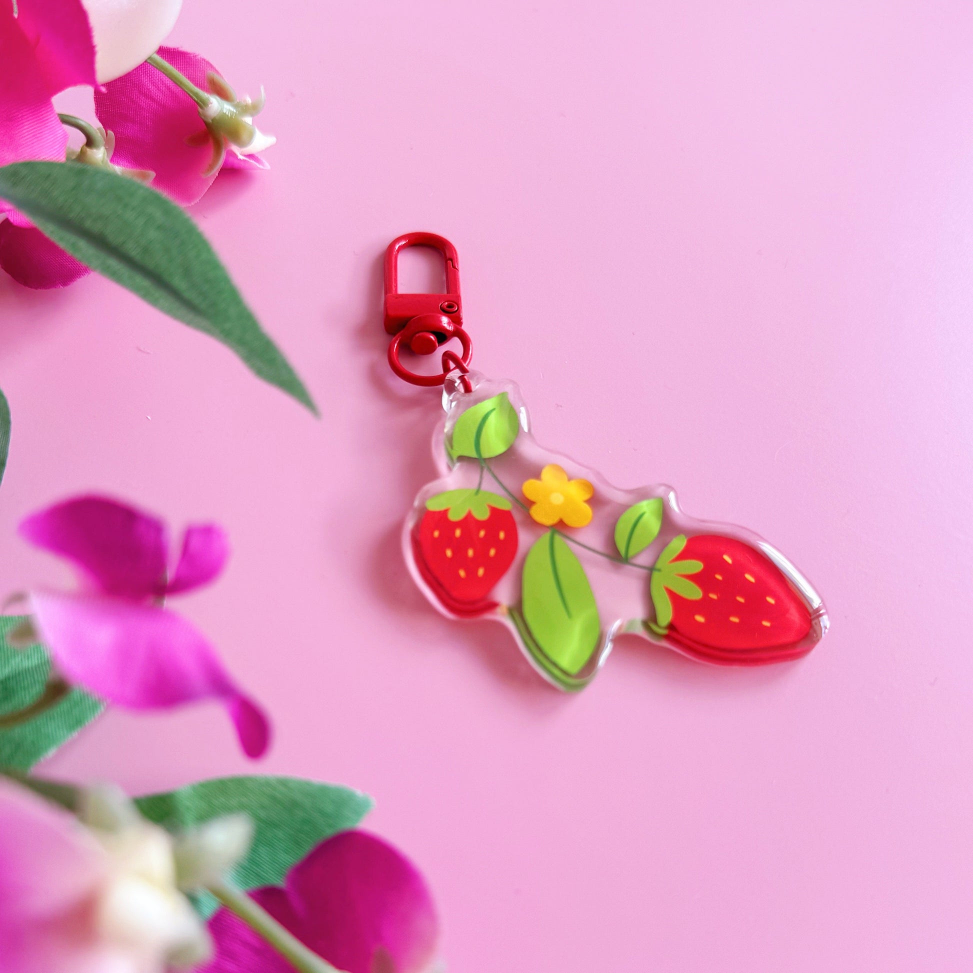 Colorful strawberry-themed keychain with a red clip on a pink background with flowers.