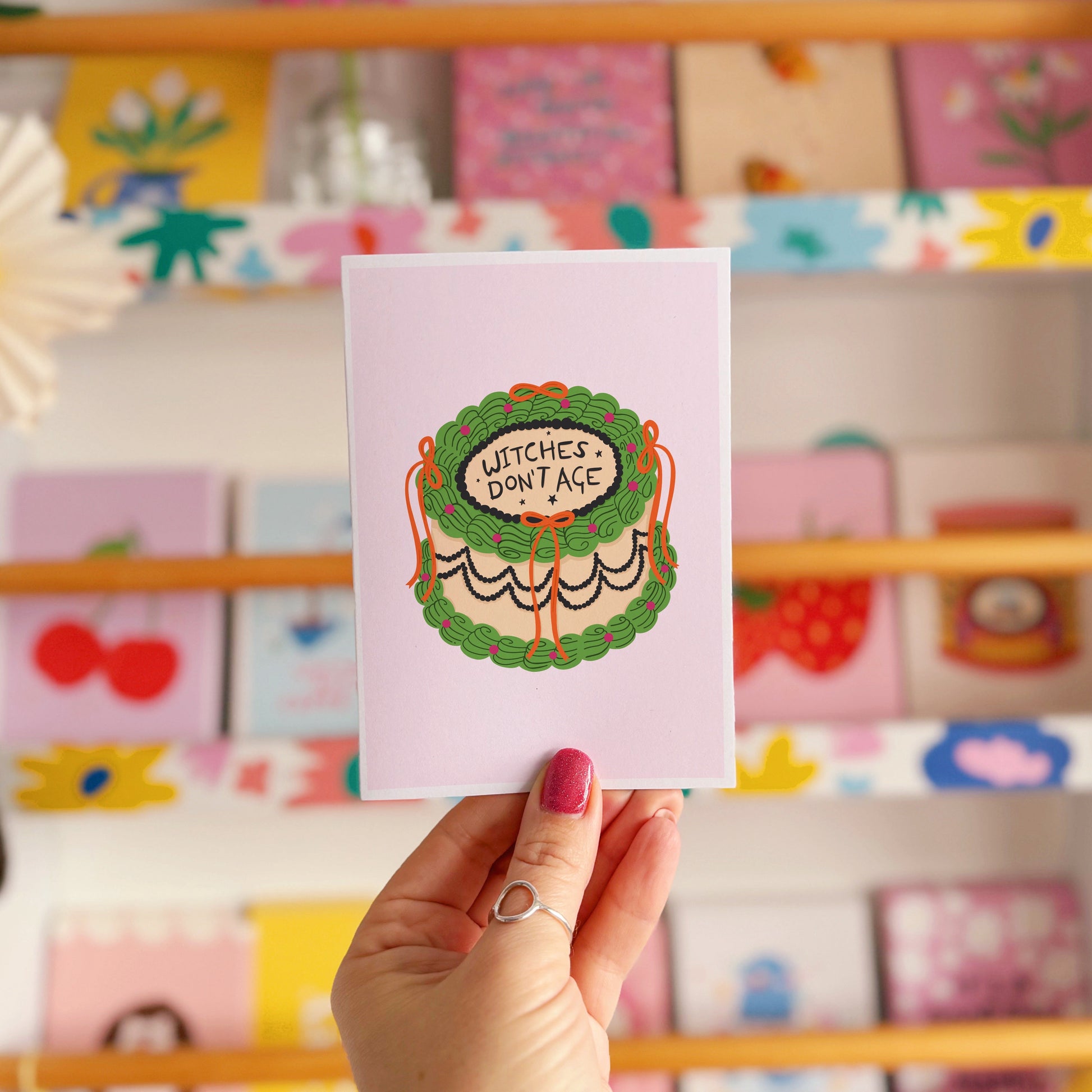 Hand holding a card with 'Witches Don't Age' design against a colourful background