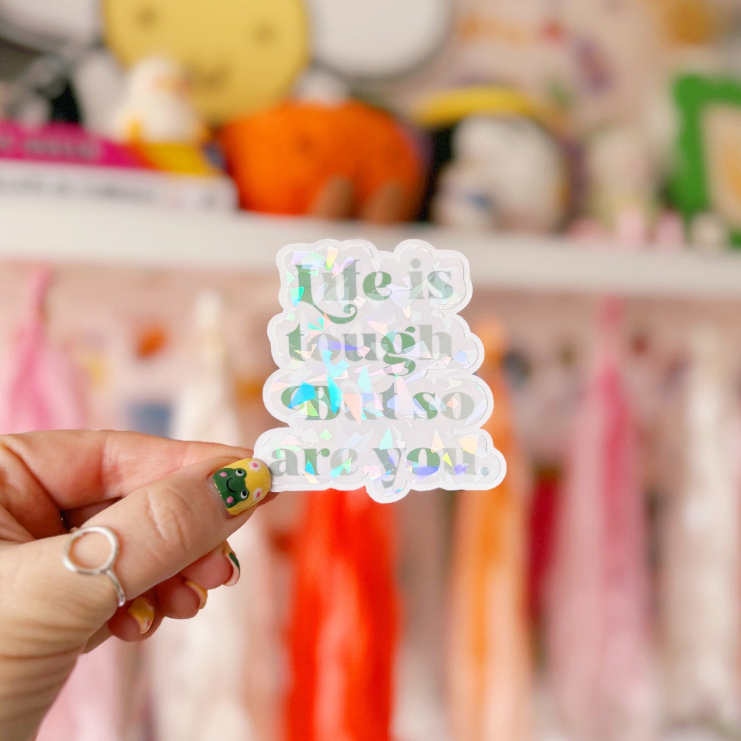 A green holographic finish sticker, with green text saying 'life is tough but so are you'.